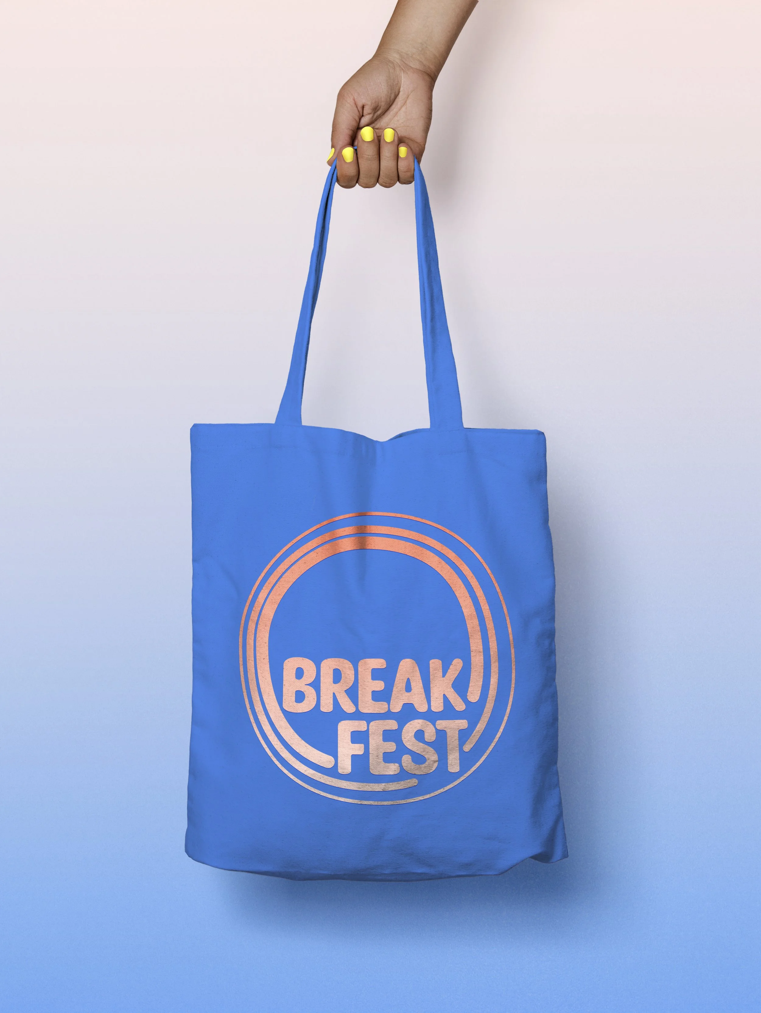 A hand with yellow nail polish holding a blue reusable tote bag with the words 'BREAK FEST' printed in pink inside a circular design.