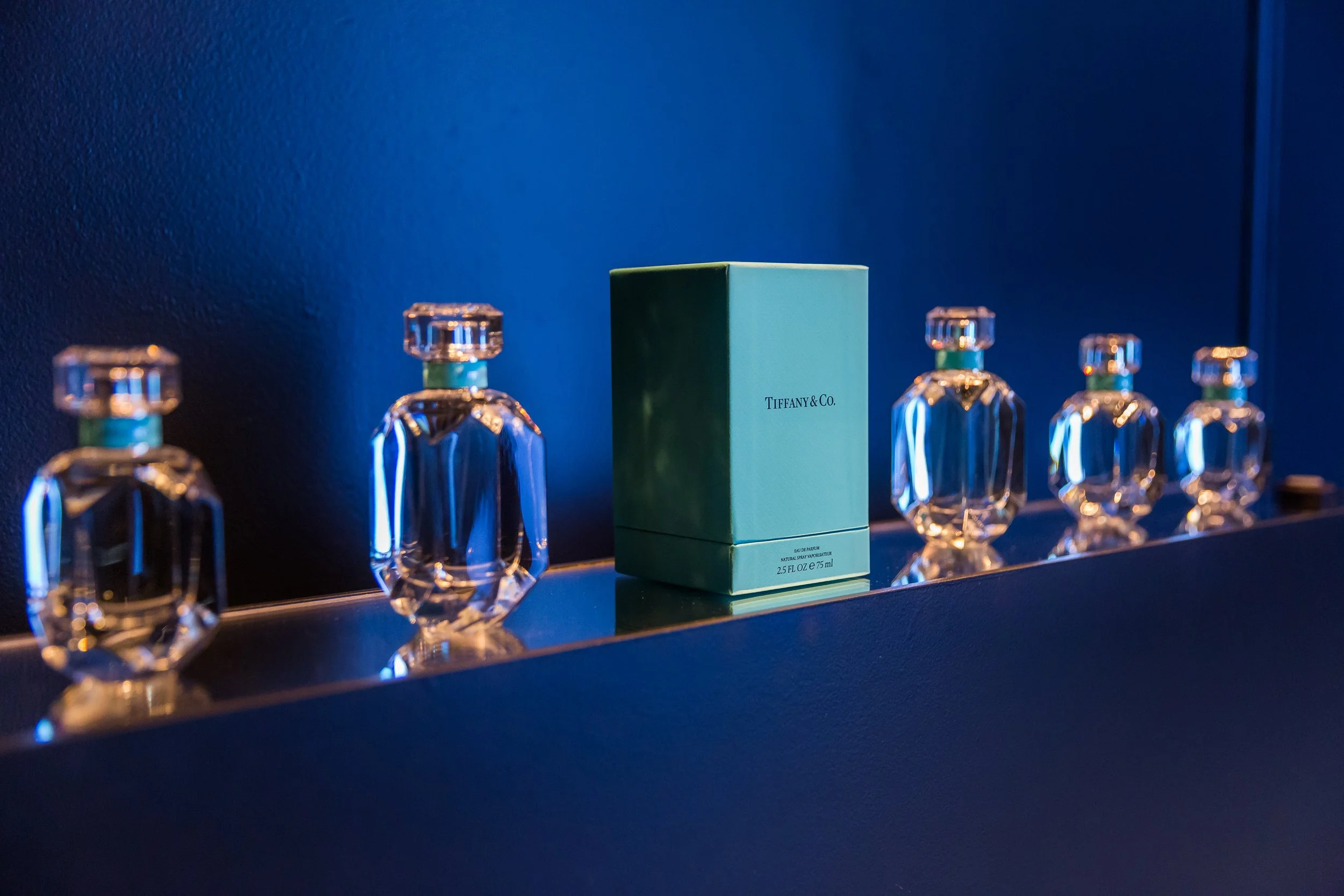 A row of five Tiffany & Co. perfume bottles with a Tiffany box in the center, displayed on a dark shelf against a dark blue wall.