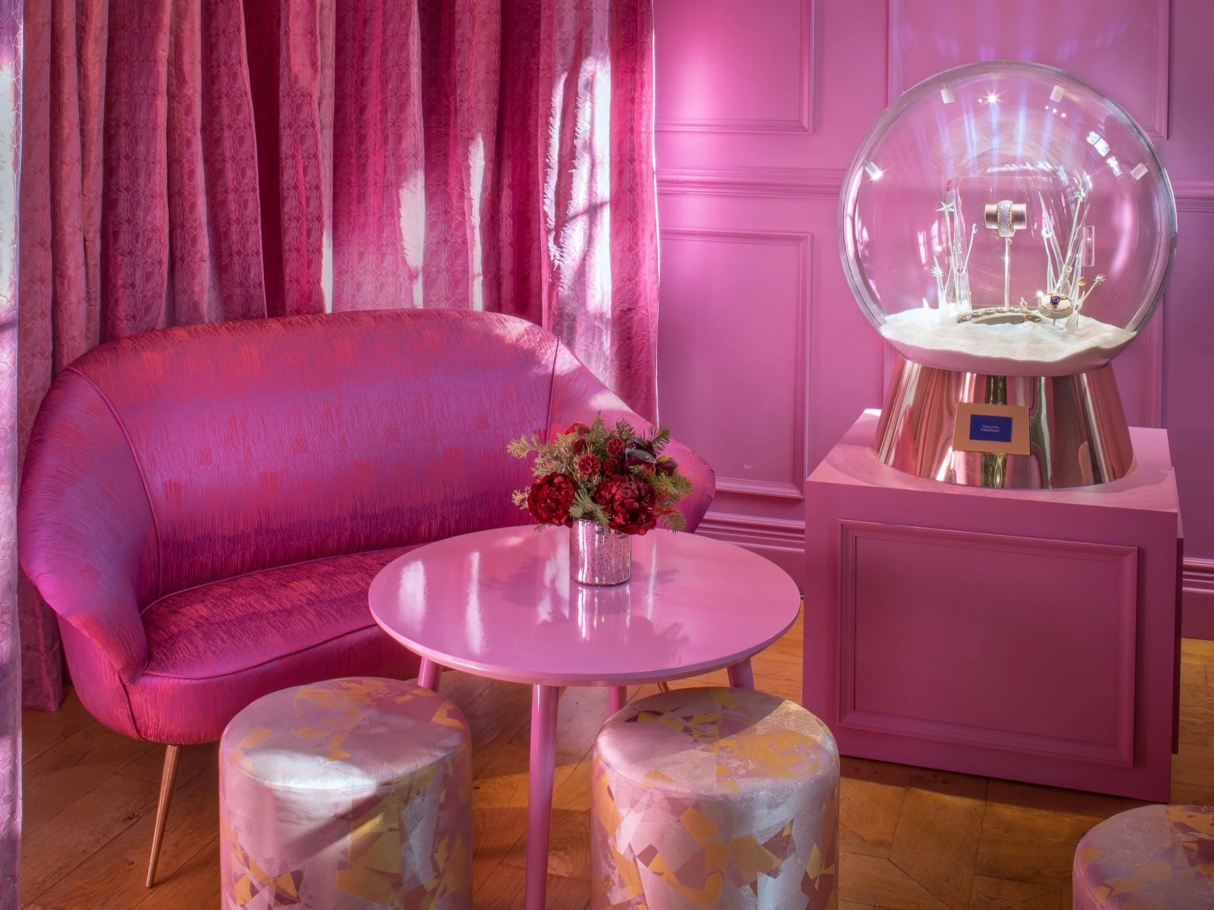 A pink velvet sofa with a flower arrangement on a small pink round table, set against pink curtains and pink paneled walls, with a glass display of jewelry on a pink stand nearby.