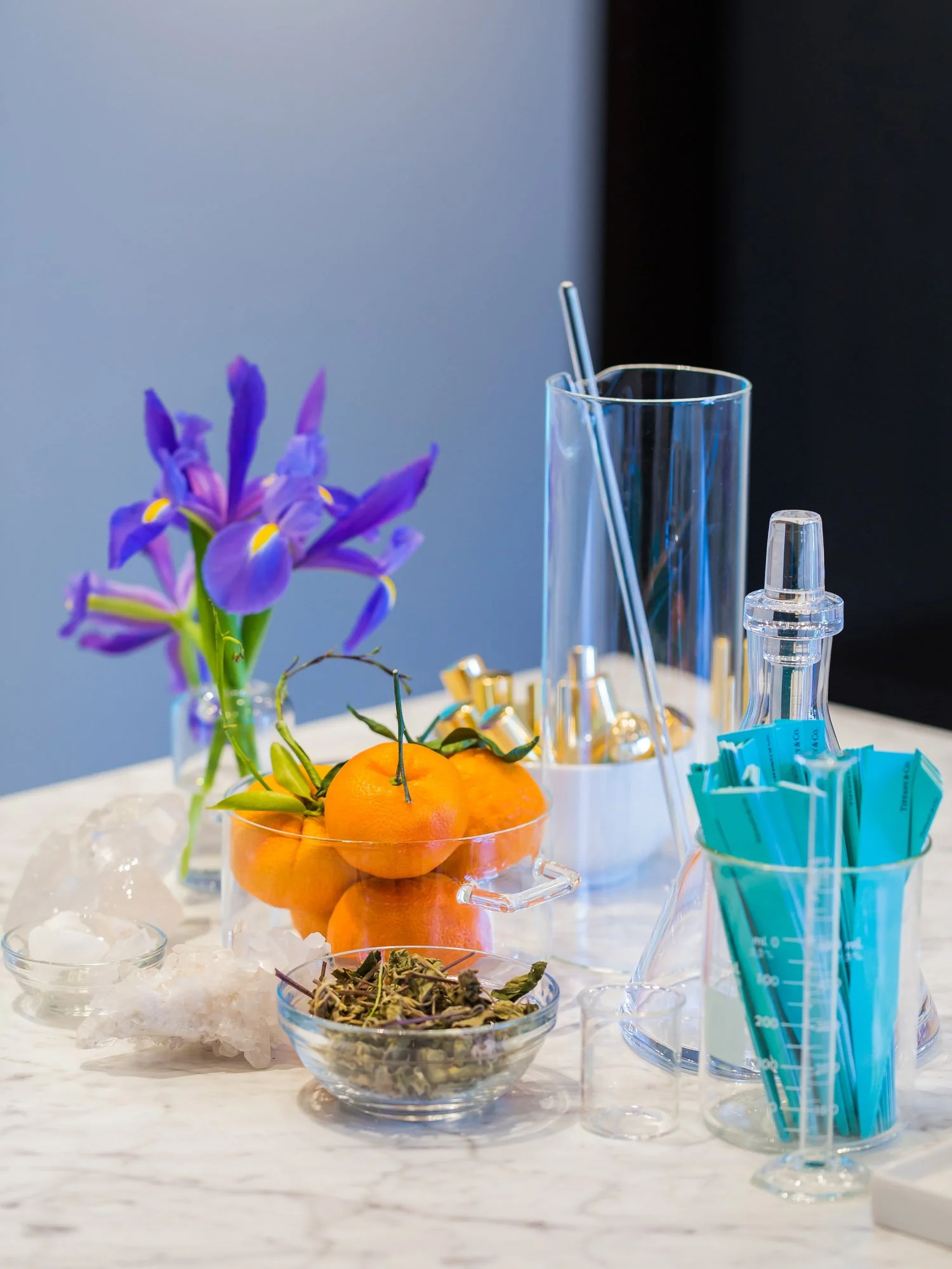 A table with a vase of purple flowers, bowl of tangerines with green leaves, various laboratory glassware, rolled-up blue paper strips, and containers of medical supplies.