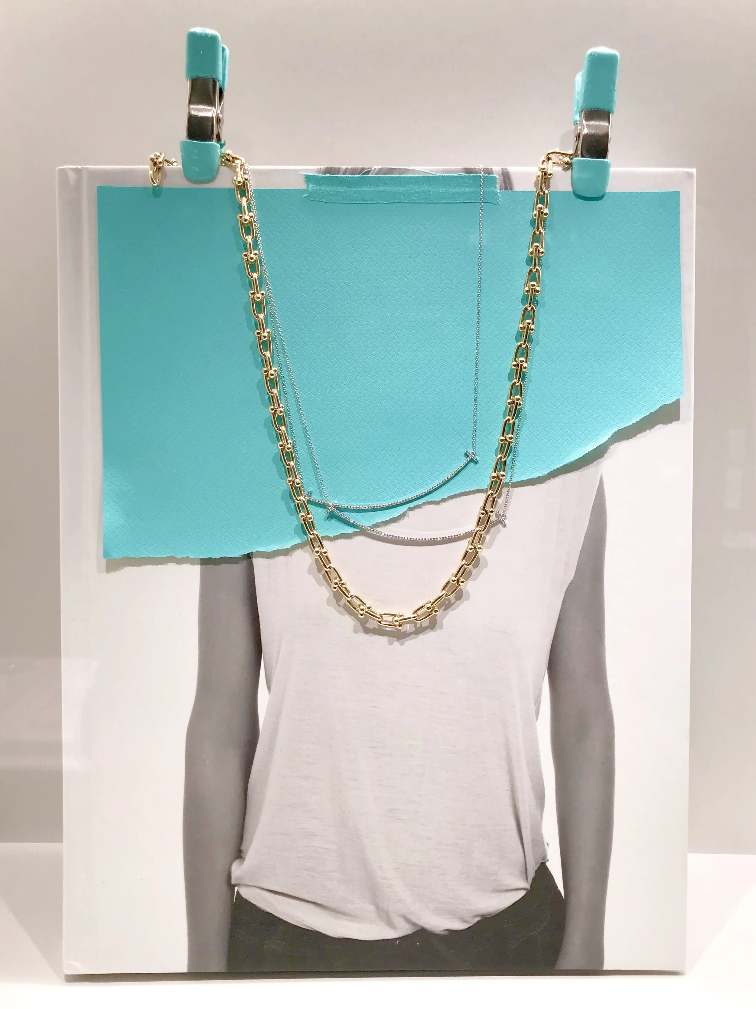 White T-shirt on a mannequin with layered gold and silver necklaces hanging from a paper sign.