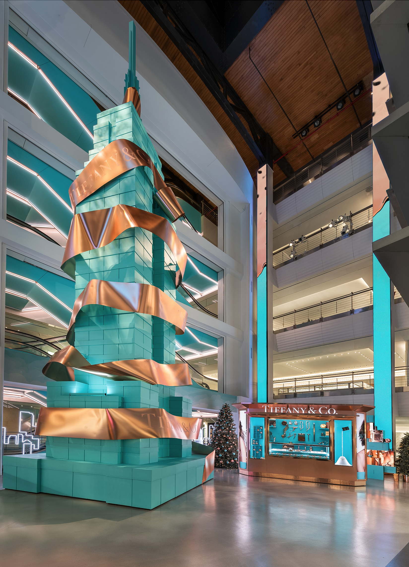 Interior of a shopping mall decorated for Christmas featuring a large turquoise display resembling a Christmas tree wrapped with wide copper-colored ribbons and a Tiffany & Co. kiosk with a Christmas tree nearby.