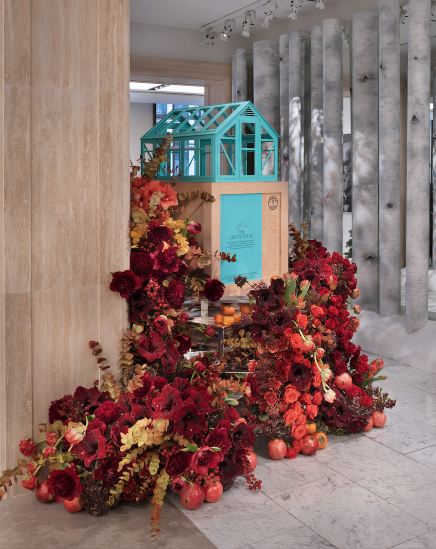 Display of a miniature greenhouse surrounded by red and burgundy flowers and pomegranates, with a sign in front titled 'The Greenhouse.'