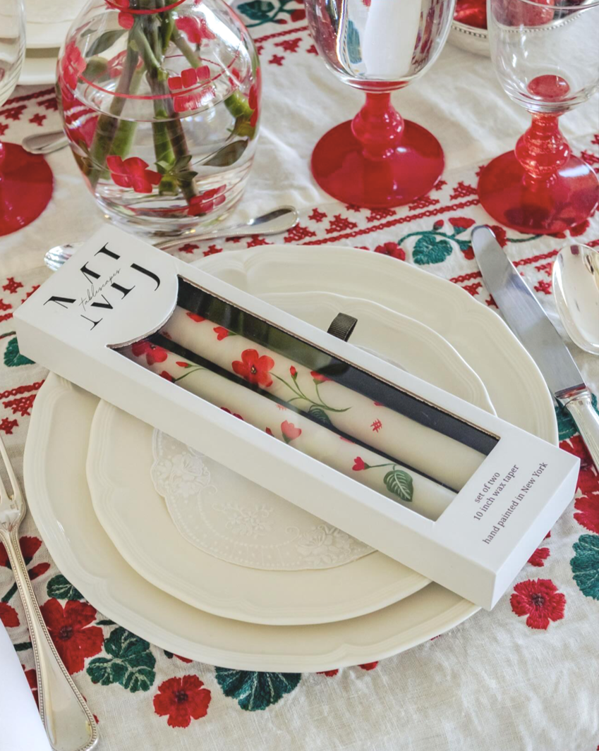 Decorative wedding or holiday table setting with red and green floral-patterned tablecloth, cream plates on a charger, and a set of white candles with a red floral design in a box, with water, flowers, and glassware.