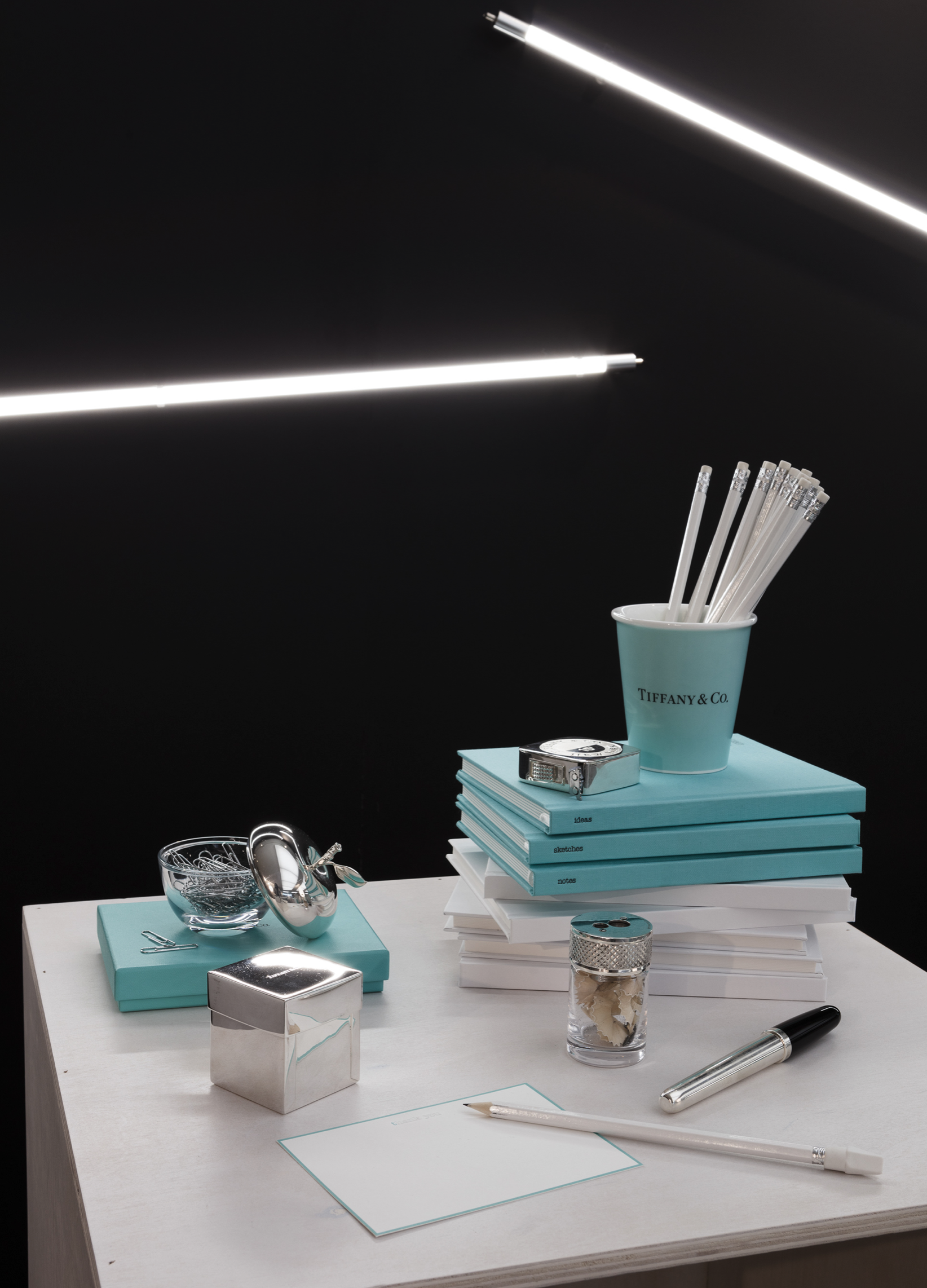 A white desk with Tiffany & Co. blue notebooks, glass bowls, a silver box, a perfume bottle, a white index card, and a silver pen. A blue cup filled with clear pens and pencils, and a silver wristwatch rest on top of the notebooks. The background is 
