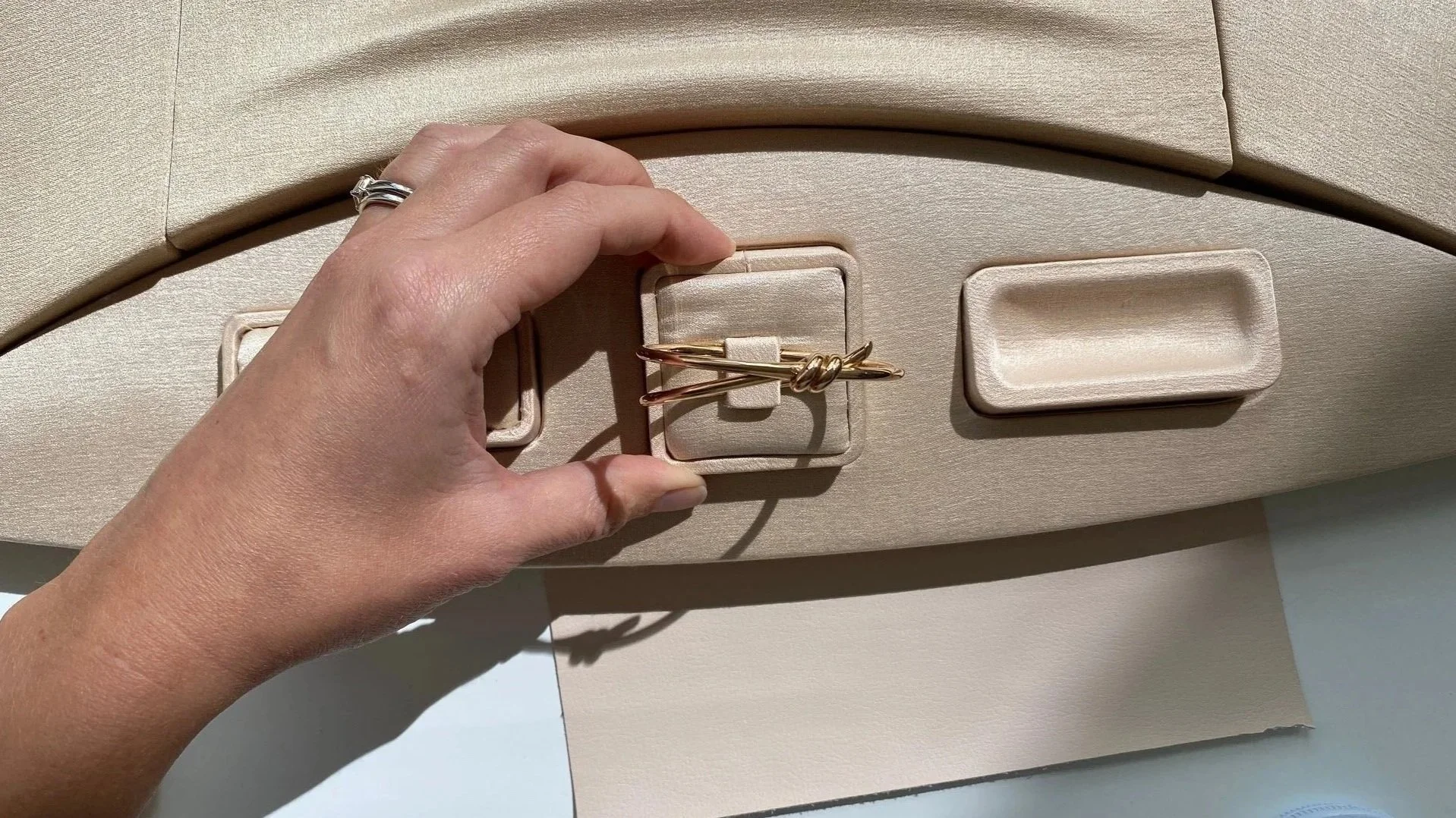 A hand arranging a gold bracelet with a knot design in a beige jewelry box surrounded by other empty jewelry compartments.