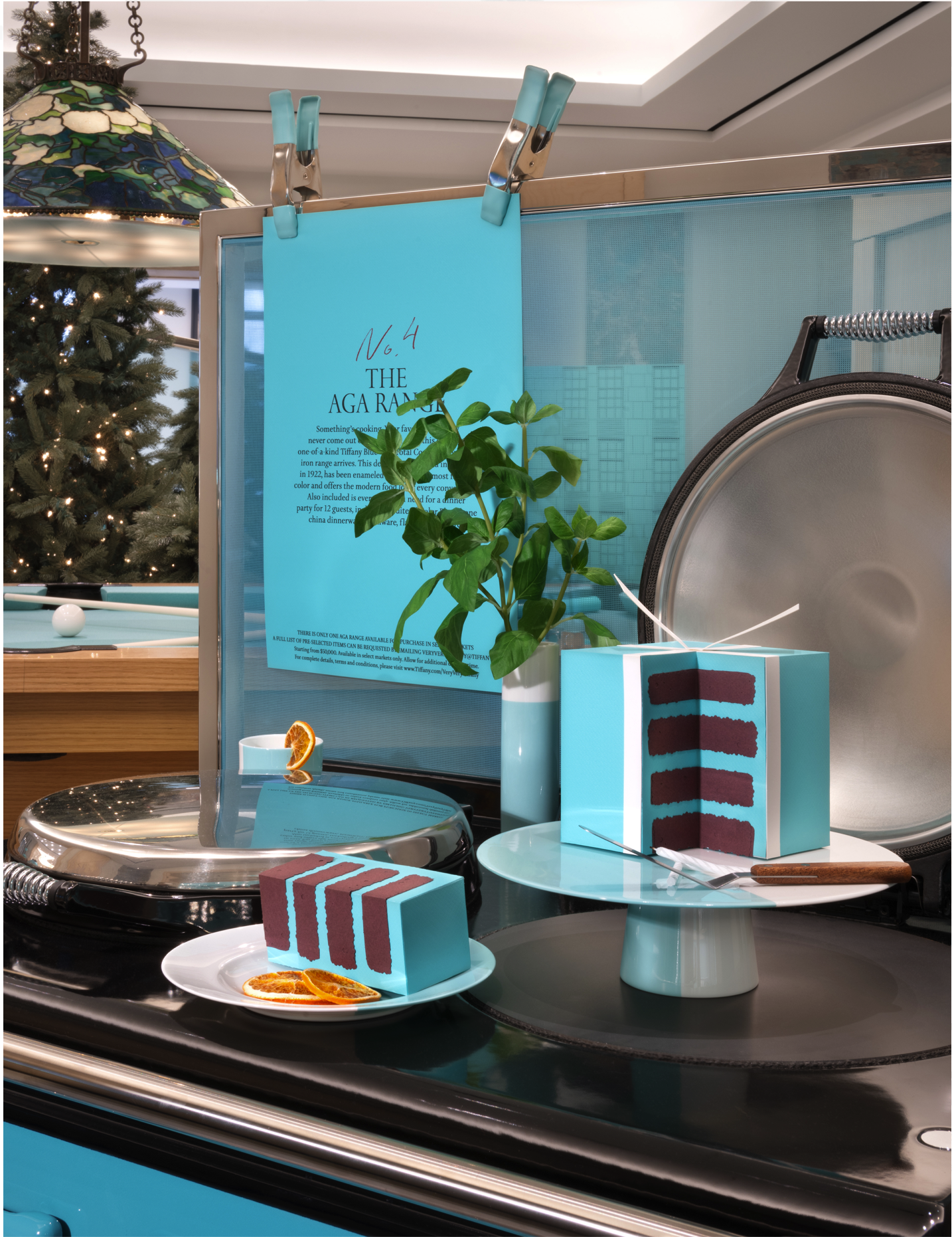 A blue cake resembling a Tiffany & Co. gift box with dark chocolate interior, placed on a cake stand. Next to it are slices of the cake on a white plate, garnished with dried orange slices. In the background, there is a green plant in a white vase, a
