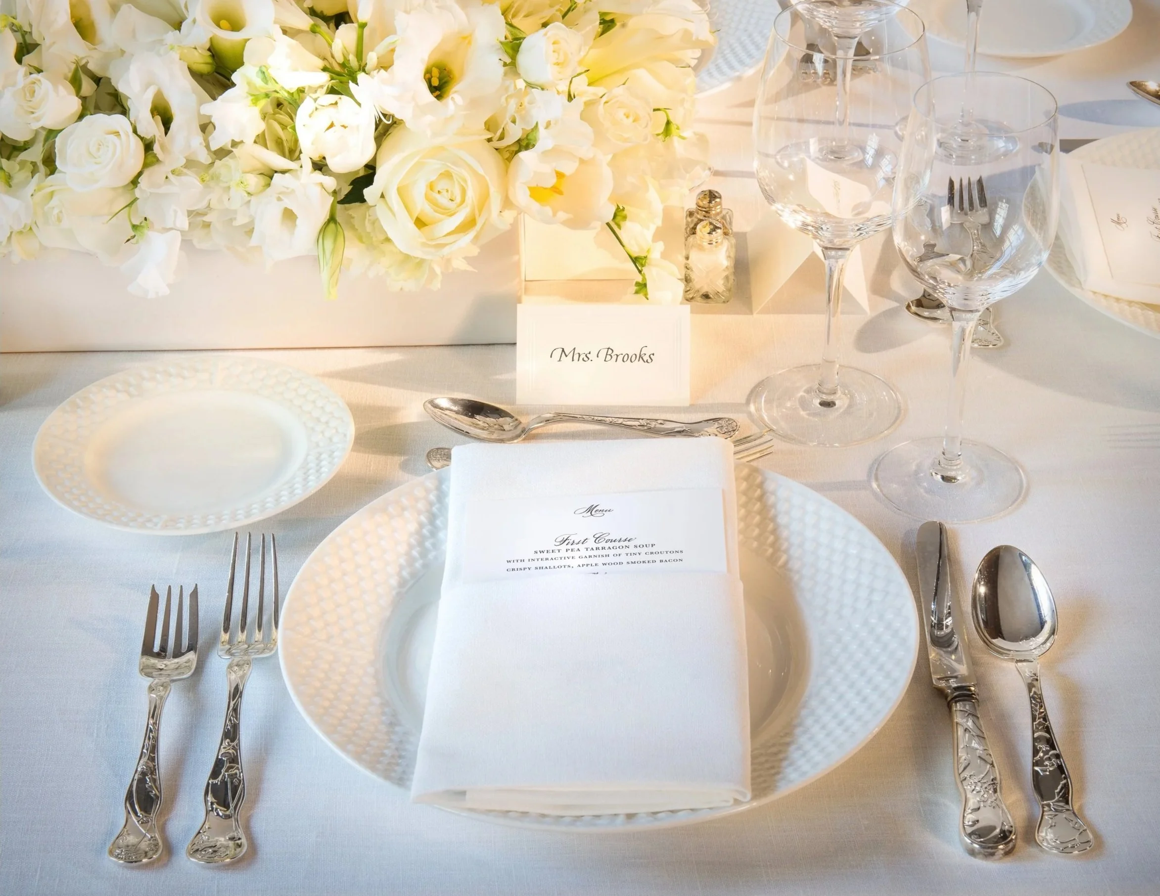 Elegant dining table set with white plates, silverware, wine glasses, a floral centerpiece of white roses and other white flowers, a place card labeled 'Mrs. Brooks,' and a menu with fine dining options.