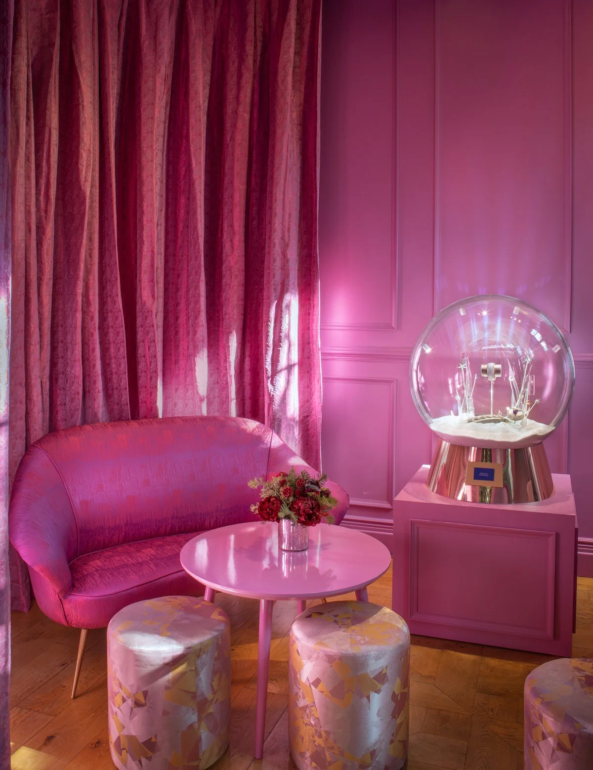 A pink-themed lounge with a pink velvet sofa, pink curtains, floral centerpiece on a pink table, and a brain exercise machine in a pink display case.