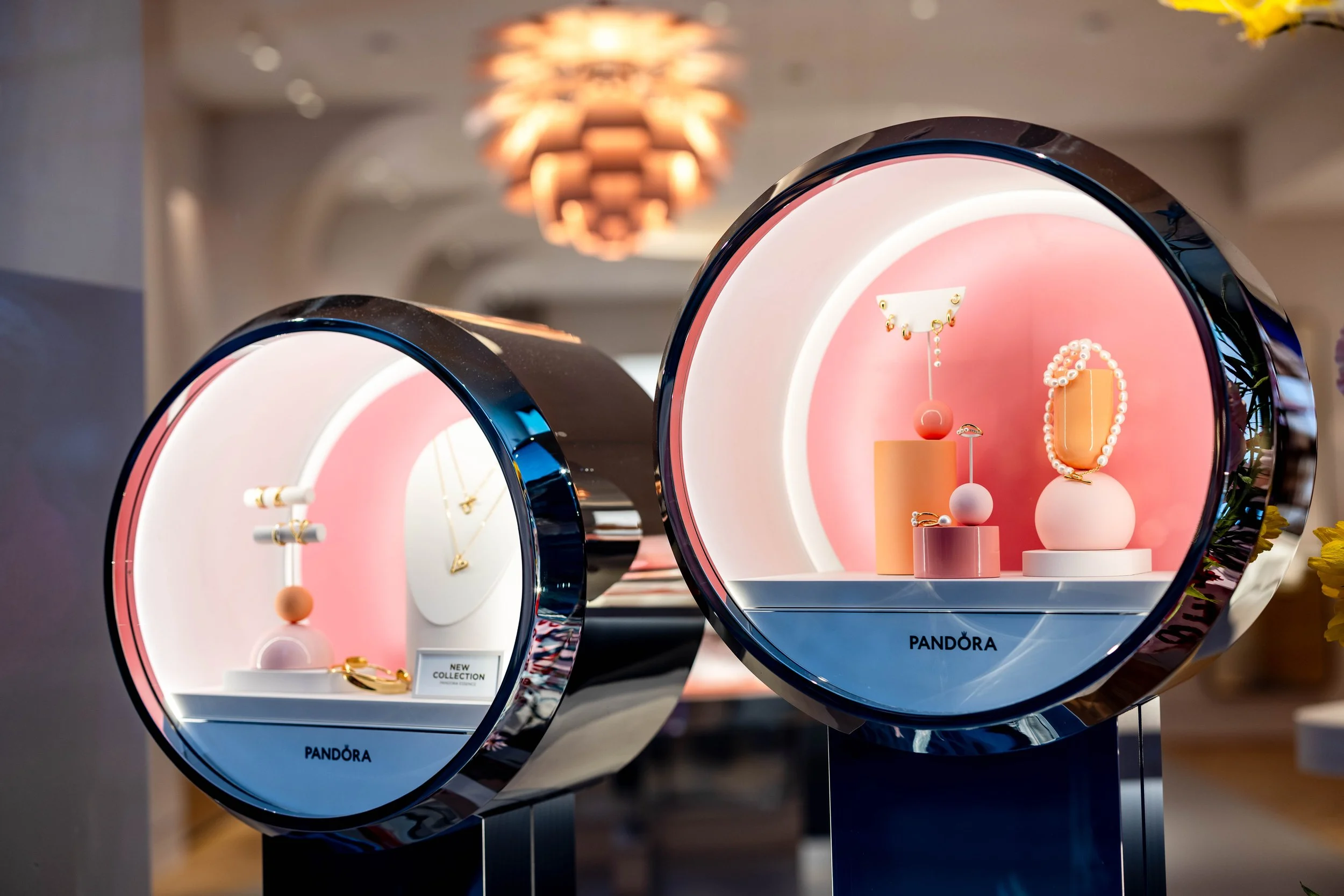 Two circular jewelry display cases with LED lighting showcase jewelry, including necklaces, bracelets, and earrings, with a pink and white background. The display case on the right has the Pandora logo at the bottom.