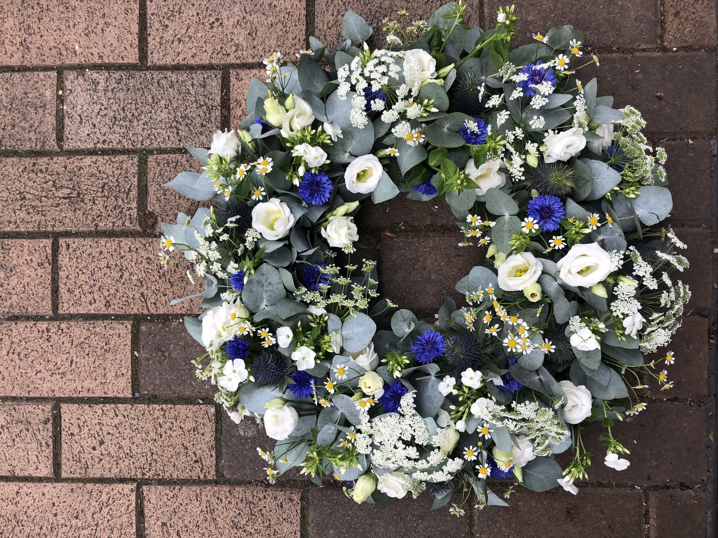 A flower arrangement in the shape of a wreath with white, blue, and green flowers and foliage, placed on a brick ground.