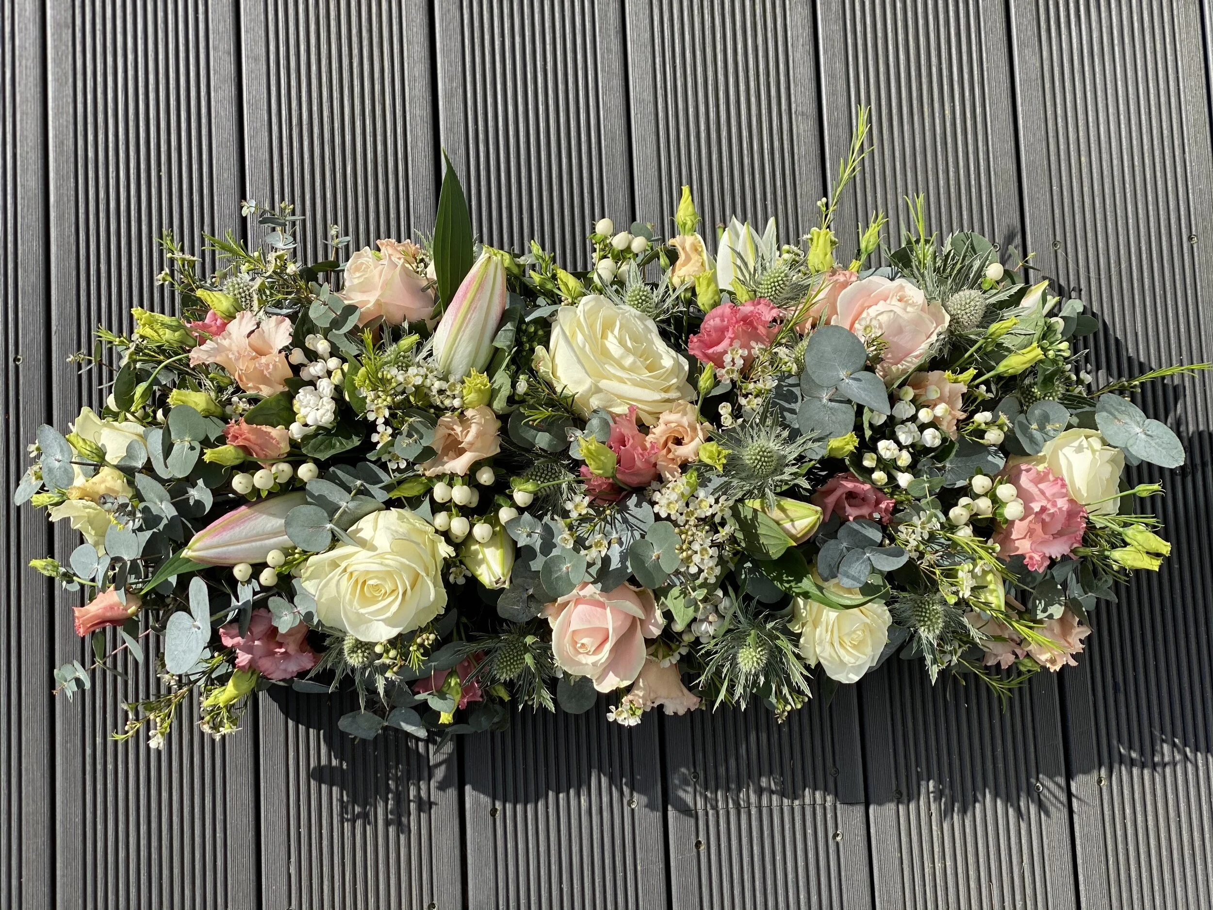 A large floral arrangement with white roses, pink lilies, pink and white other flowers, green leaves, and eucalyptus, placed on a dark wooden surface.