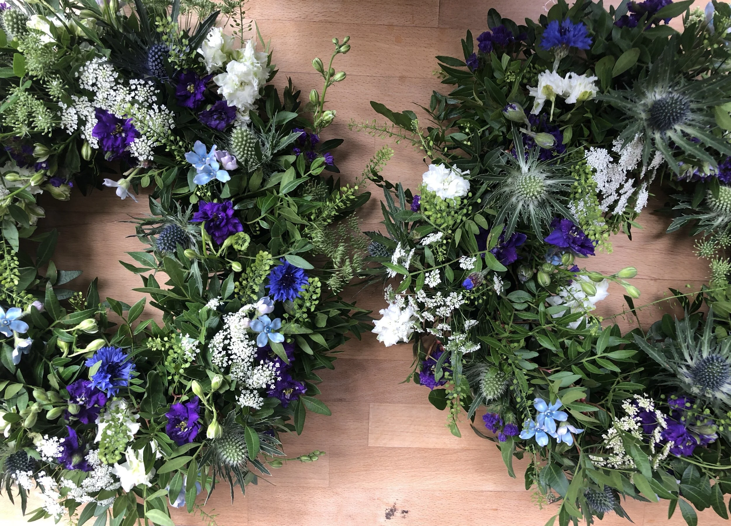 Two floral wreaths with greenery, purple, white, and light blue flowers, arranged on a wooden floor.