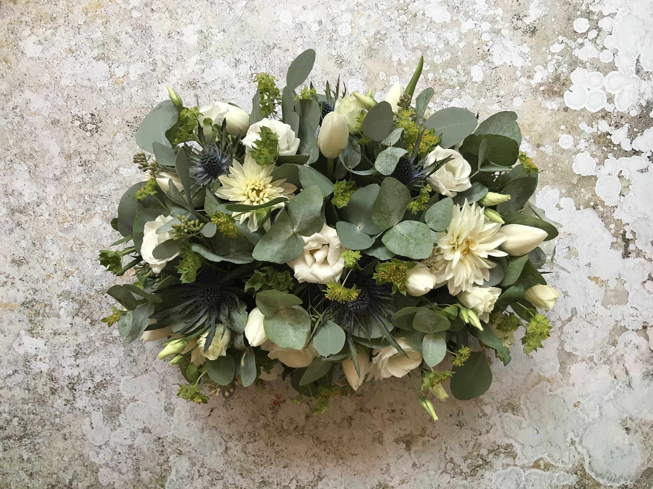 White floral bouquet with assorted white flowers, green foliage, and grayish-green leaves on a textured, weathered light-colored surface.