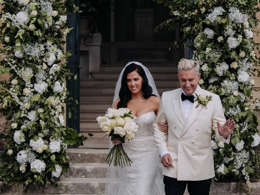 A bride in a white wedding dress holding a bouquet of white roses stands next to a groom in a white tuxedo jacket with a black bow tie, smiling and walking down stone steps decorated with lush white and green floral arrangements.