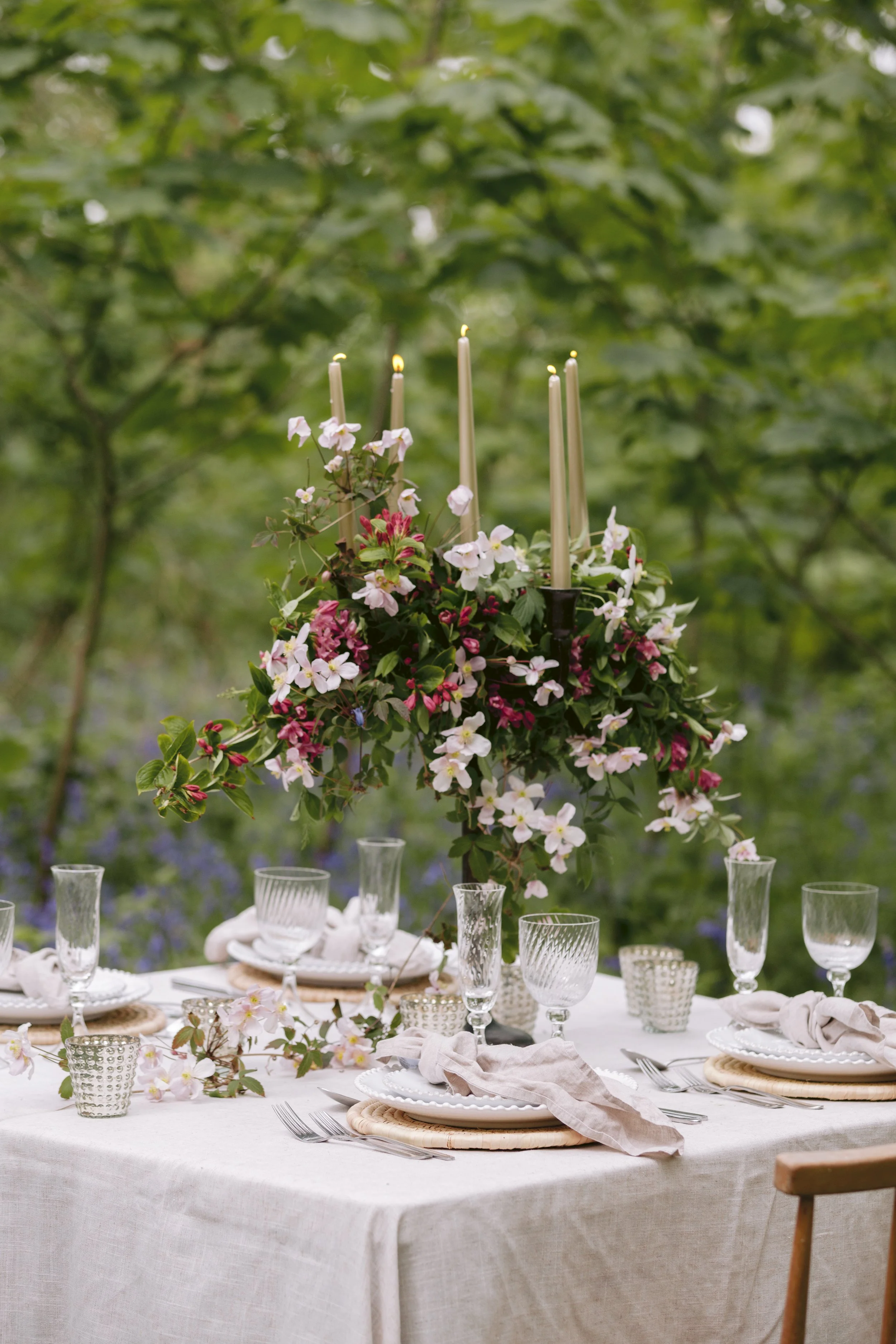 Elegant outdoor table setting with a floral centerpiece and candles, surrounded by greenery.