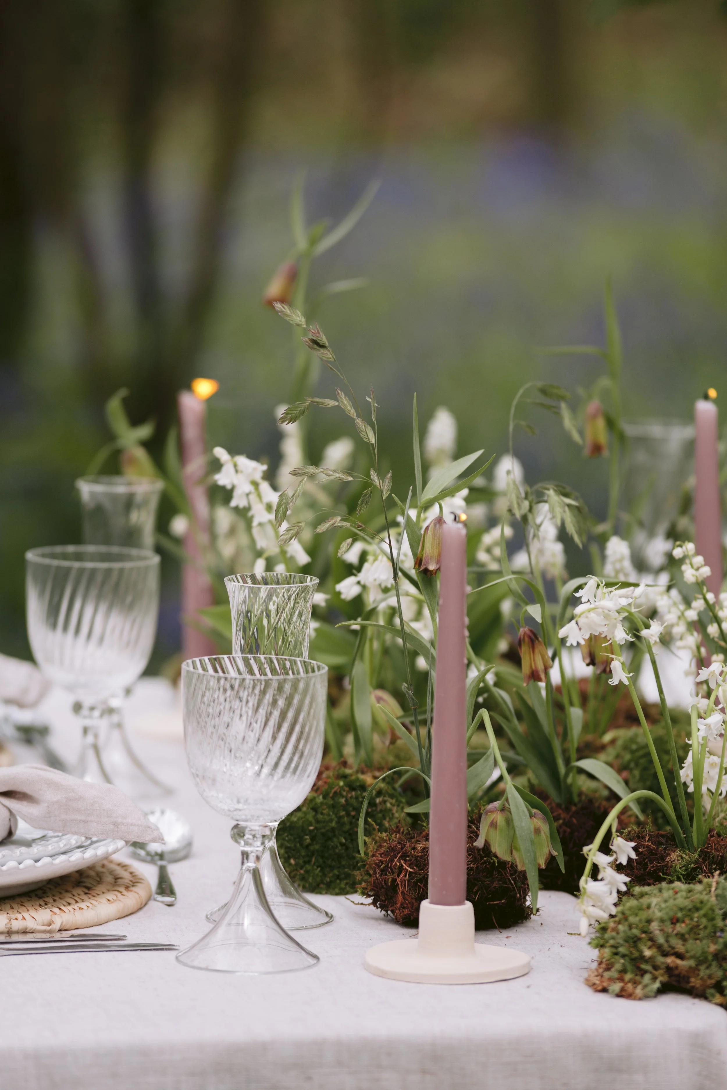 Table setting with clear patterned glasses, pink candles, white flowers, and greenery in an outdoor garden setting.