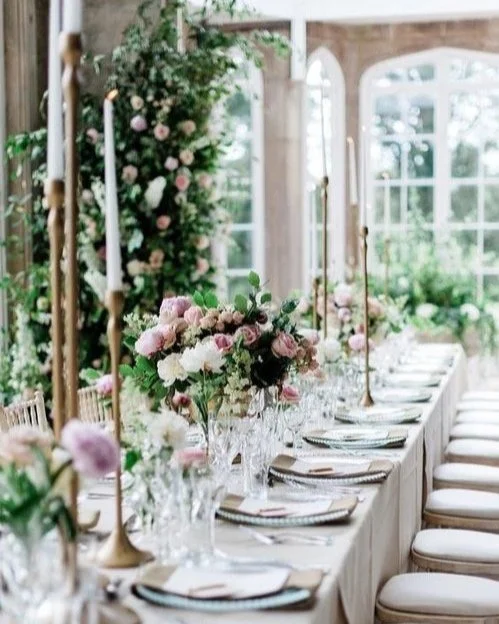 Elegant indoor dining table decorated with pink and white flowers, tall gold candlesticks, and white napkins, with large windows and greenery in the background.