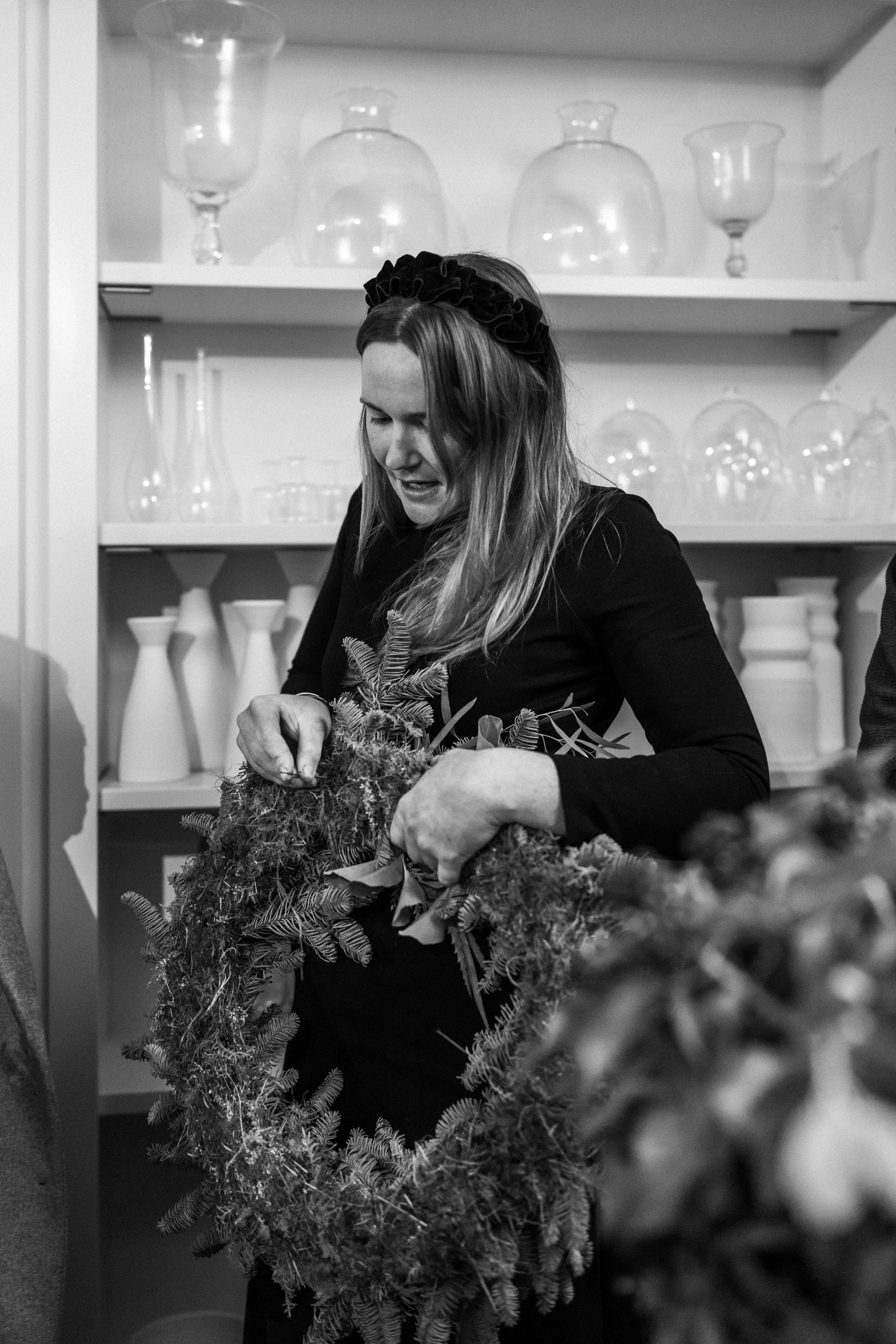 A woman is working on a Christmas wreath, surrounded by glass vases and jars on shelves in the background.