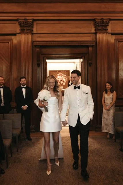 Bride in a white dress holding a bouquet walking down the aisle with groom in a tuxedo in a wood-paneled room.