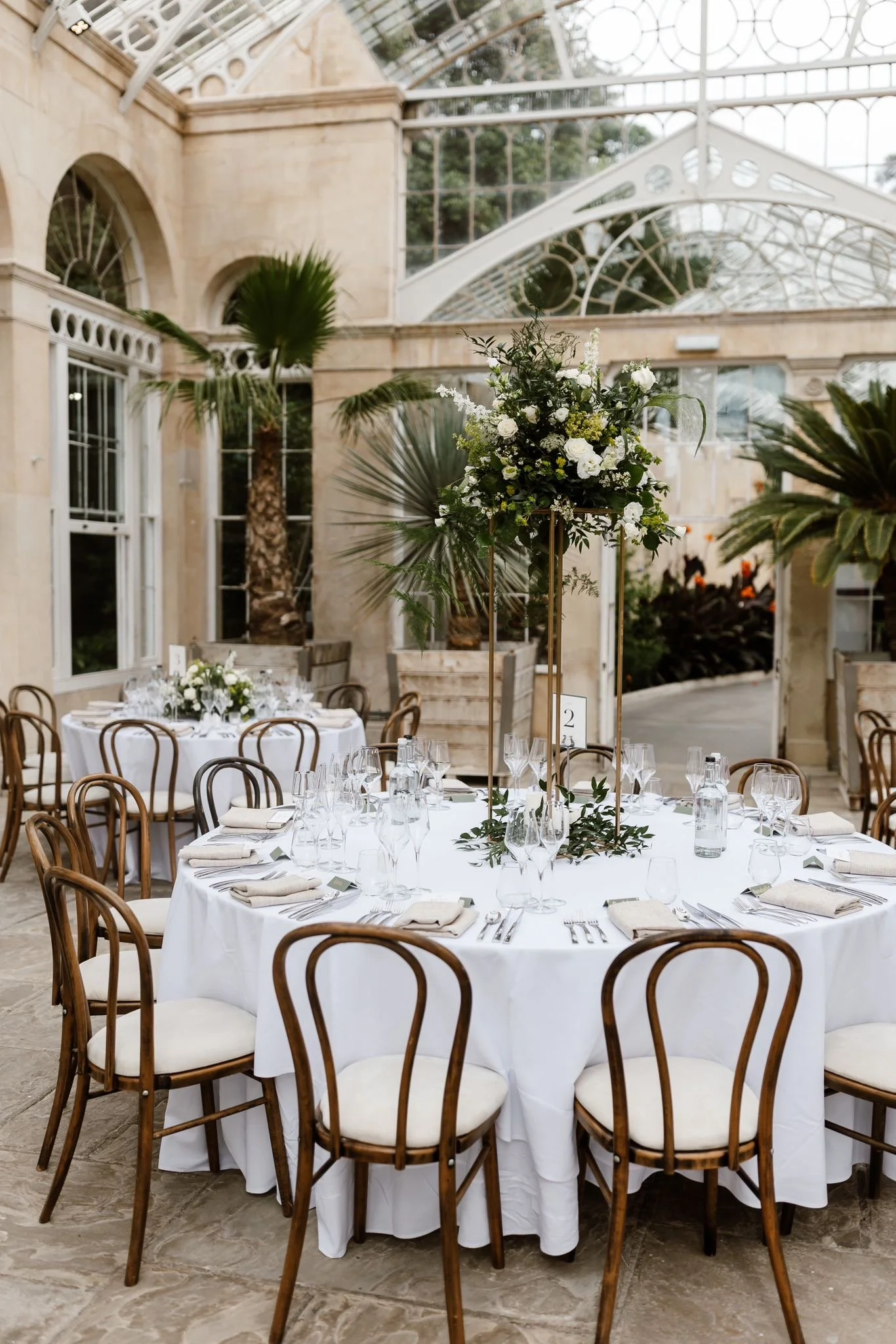 Elegant round banquet table set for a formal event inside a glass conservatory with a lush tropical plant backdrop, featuring wooden chairs, white tablecloth, glassware, and floral centerpiece.