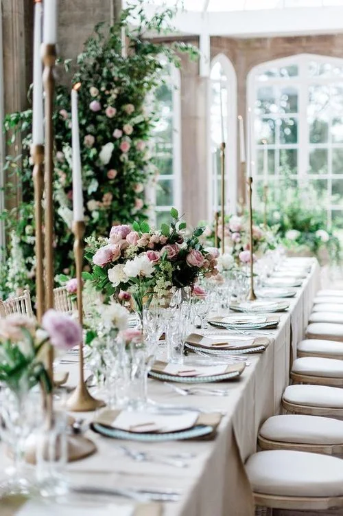 Long dining table decorated for a formal event with floral centerpieces, tall candles, and elegantly set plates and glasses, in a bright room with large windows.
