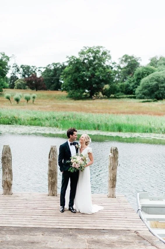 A wedding couple standing on a wooden dock by a lake, with a bride holding a bouquet, and trees and grassy fields in the background.