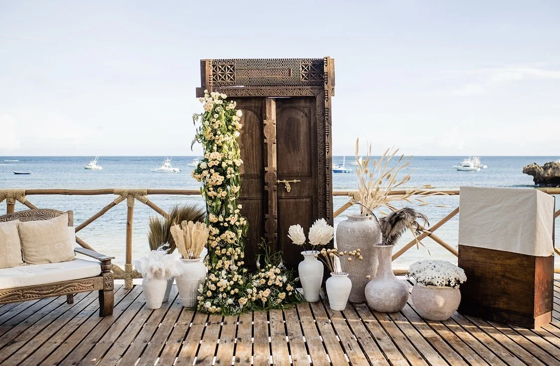 Beachside wedding setup with a wooden door, floral decorations, vases, a bench, and a table on a wooden deck overlooking the ocean with boats.