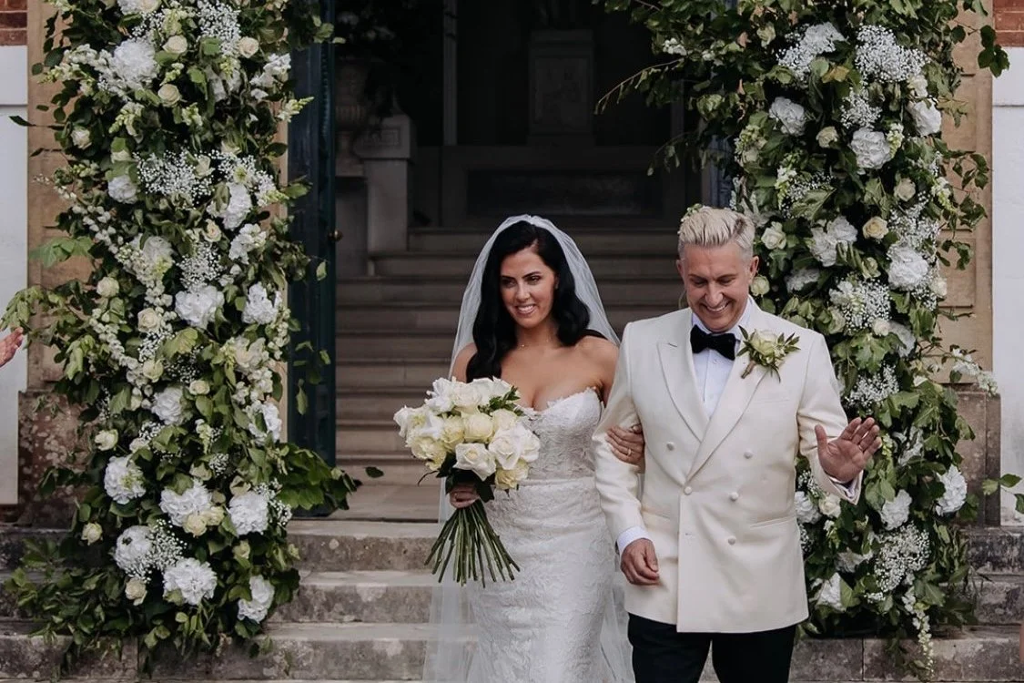 A bride with dark hair in a white wedding dress holding a bouquet, walking down stairs with an older man in a white tuxedo with black bowtie, surrounded by large floral arrangements on either side of the stairway.