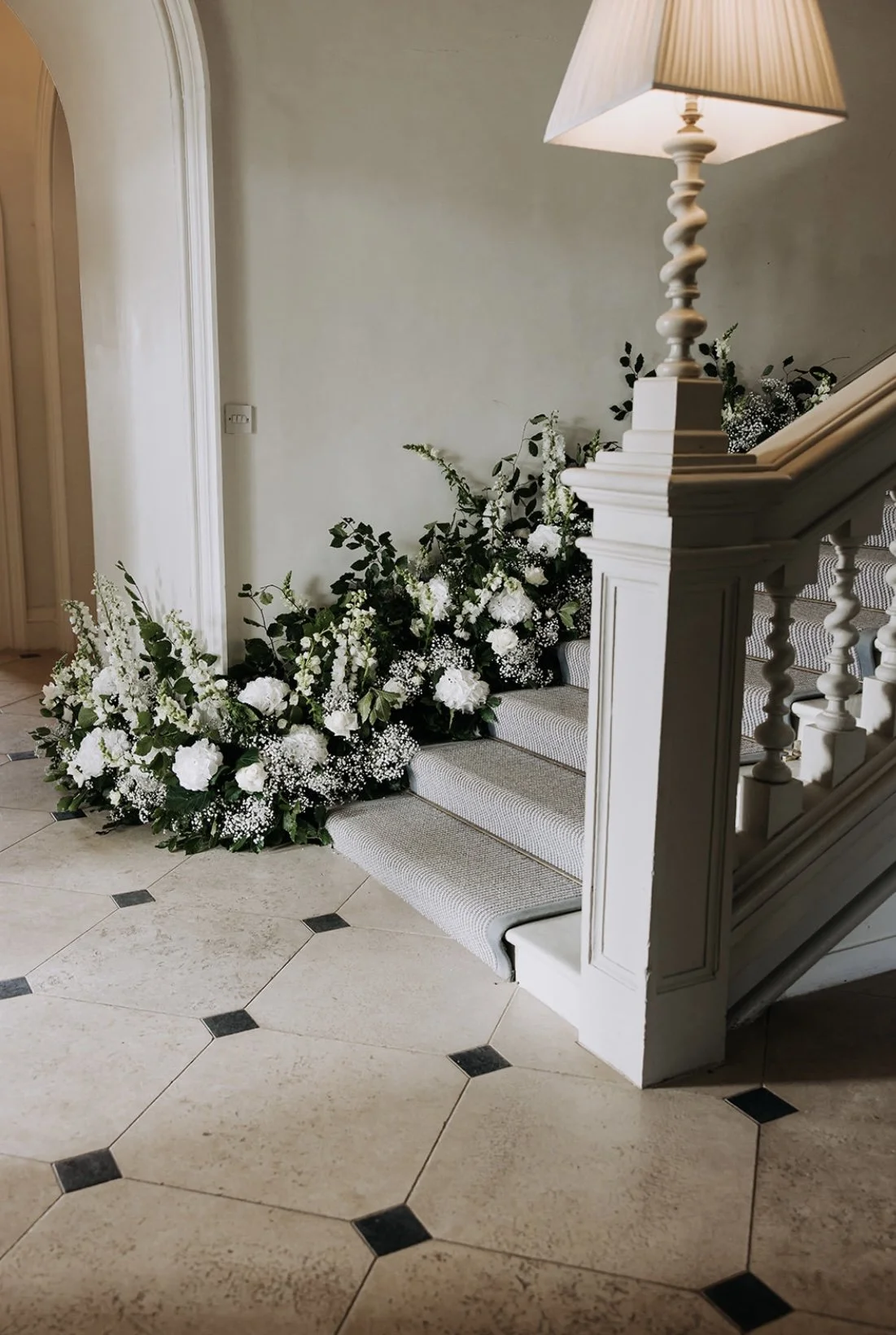 A staircase decorated with white flowers and greenery, with a lamp on the landing, in an indoor setting with tiled flooring.
