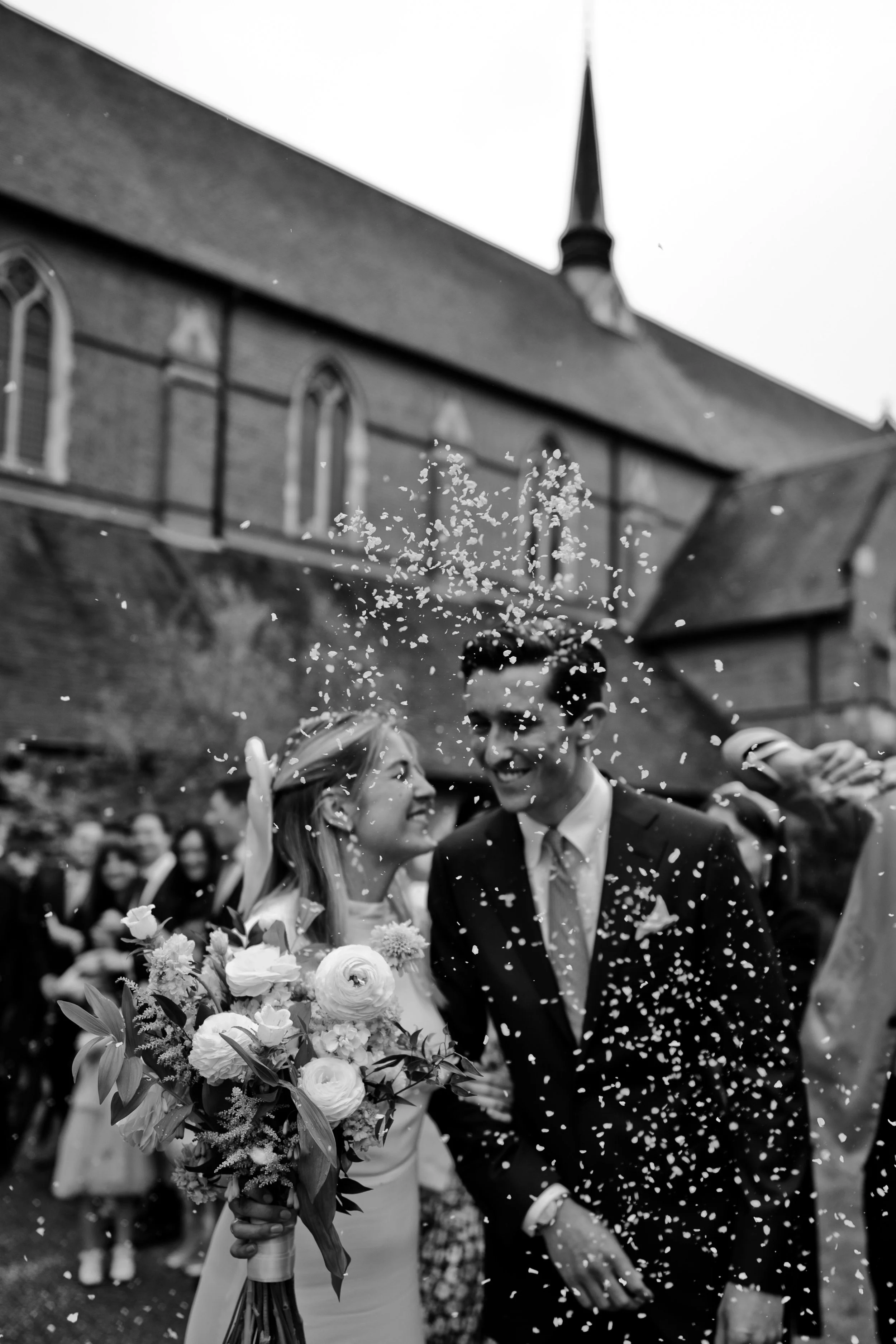 A black and white photo of a newlywed couple celebrating outdoors, with confetti falling around them, holding a bouquet of flowers, in front of a church.