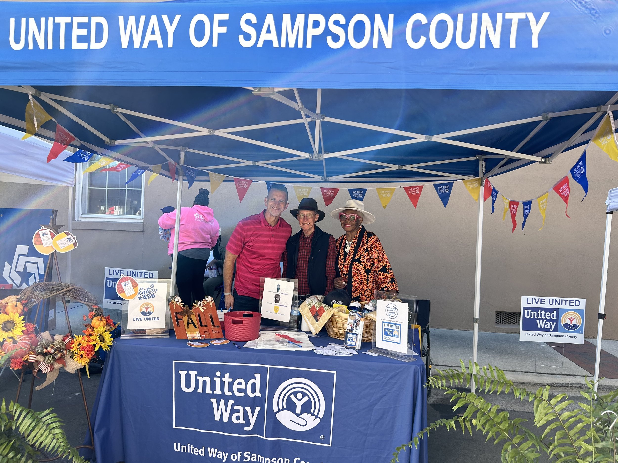 Blue pop up event tent with "UNITED WAY OF SAMPSON COUNTY" across the top of image. Three people stand behind a branded table, a tall middle aged white man on left, an older white gentleman in the middle, and an older black lady on the right.