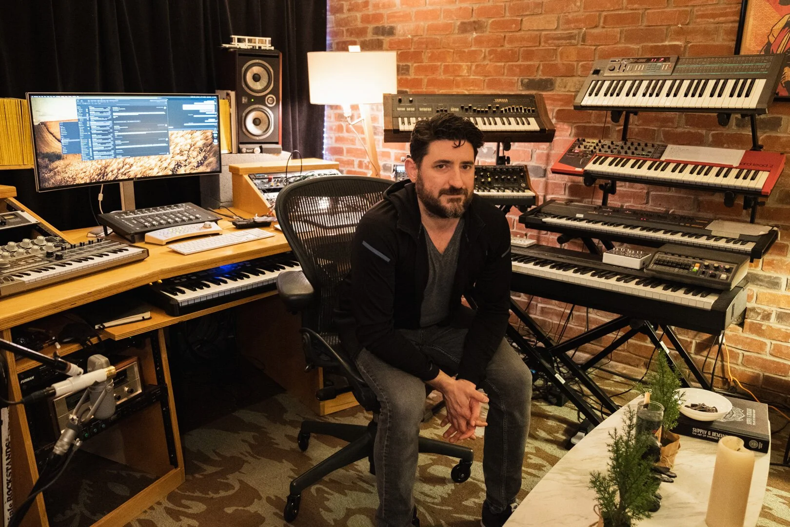 A man with dark hair and a beard sitting in a music studio surrounded by keyboard synthesizers, a computer monitor, studio speakers, and recording equipment, with a brick wall in the background.