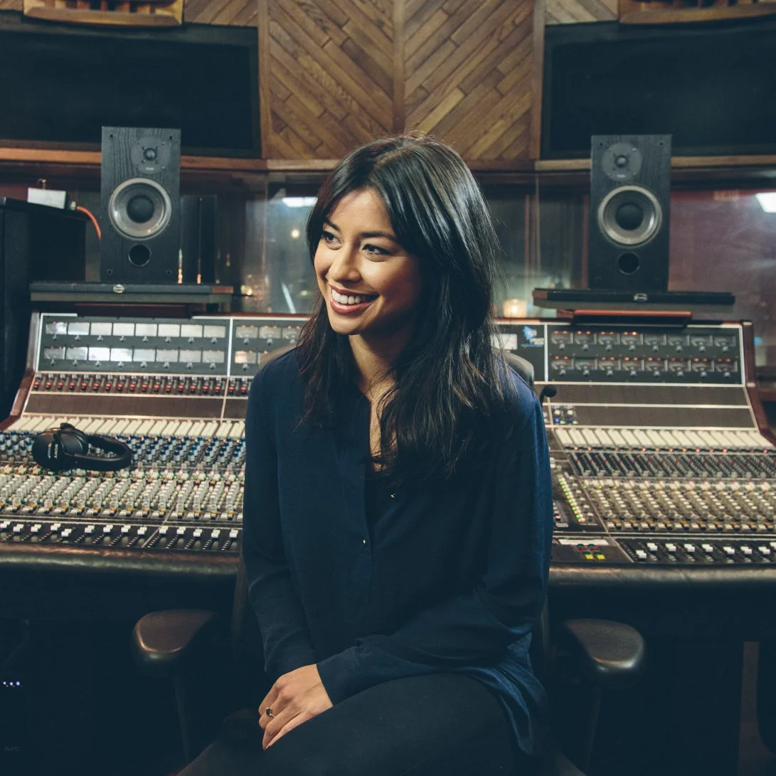 A woman sitting in a recording studio with professional audio mixing equipment and speakers behind her.