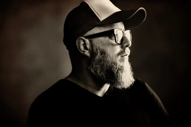 Profile of a man with a beard wearing glasses and a baseball cap, in black and white.