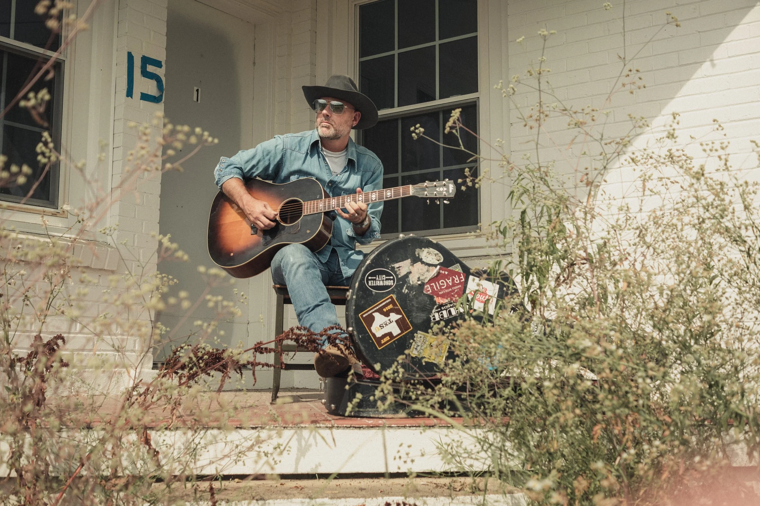 A man sitting on a porch playing an acoustic guitar, wearing sunglasses, a wide-brimmed hat, a denim jacket, and jeans, with a guitar case in front of him, surrounded by plants and bushes.