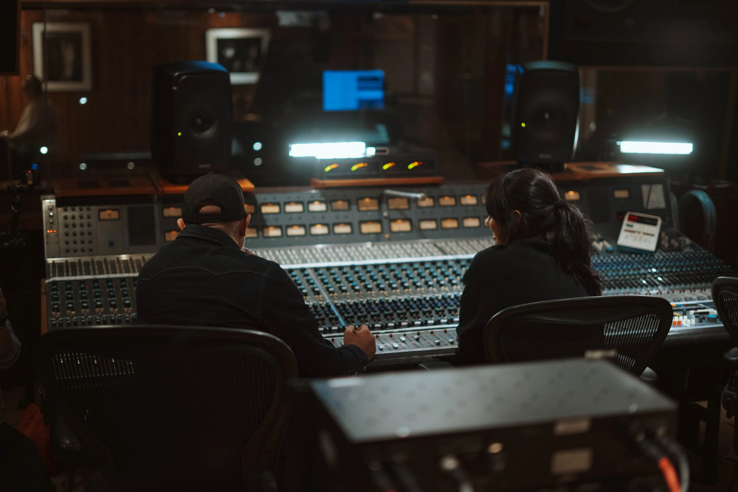 Two people sit at a large audio mixing console in a recording studio, with black speakers and computer monitors in the background.