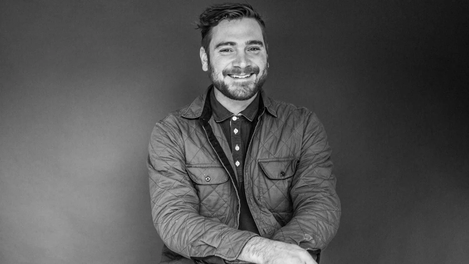 Black and white photo of a smiling man with a beard and short hair, wearing a quilted jacket over a shirt, sitting with his arms crossed against a plain background.