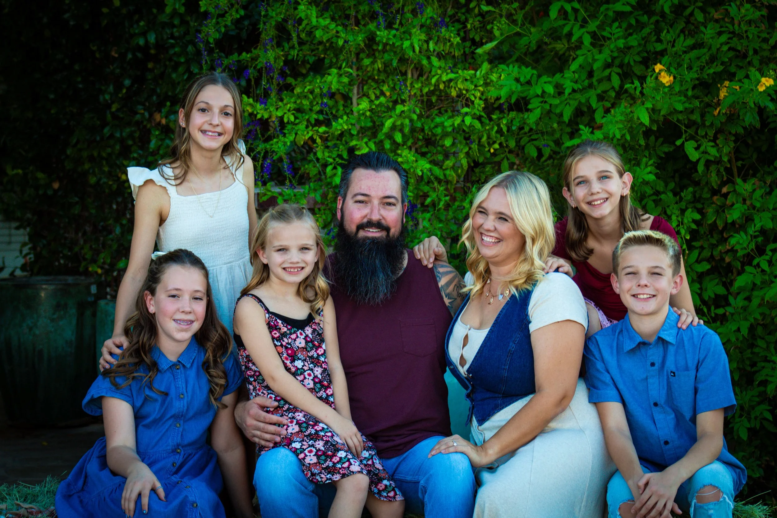 A group photo of a family of nine people, including adults and children, smiling and posing outdoors with greenery in the background.
