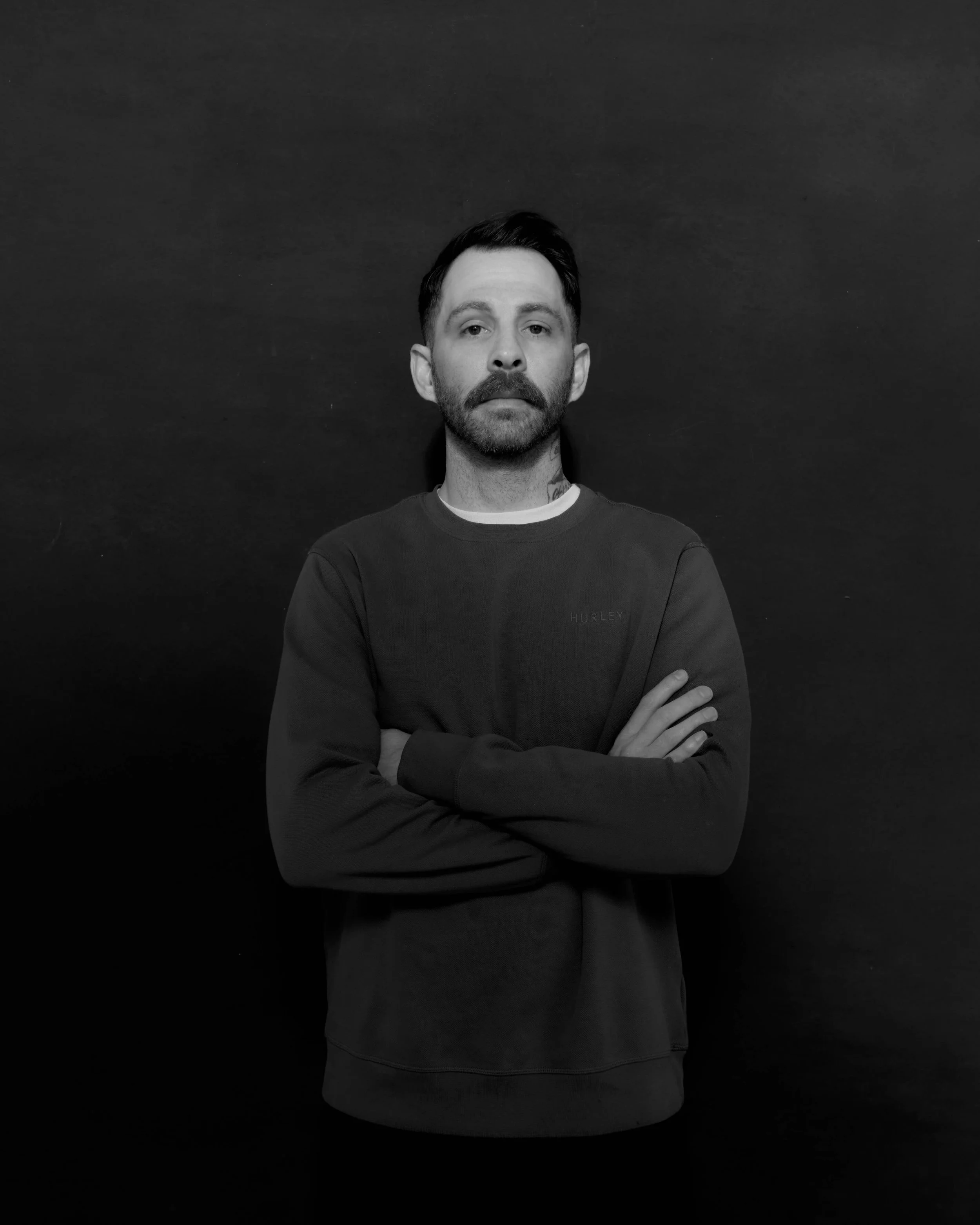A black-and-white portrait of a man with short dark hair, a beard, and mustache, wearing a dark sweatshirt with crossed arms, standing against a dark background.
