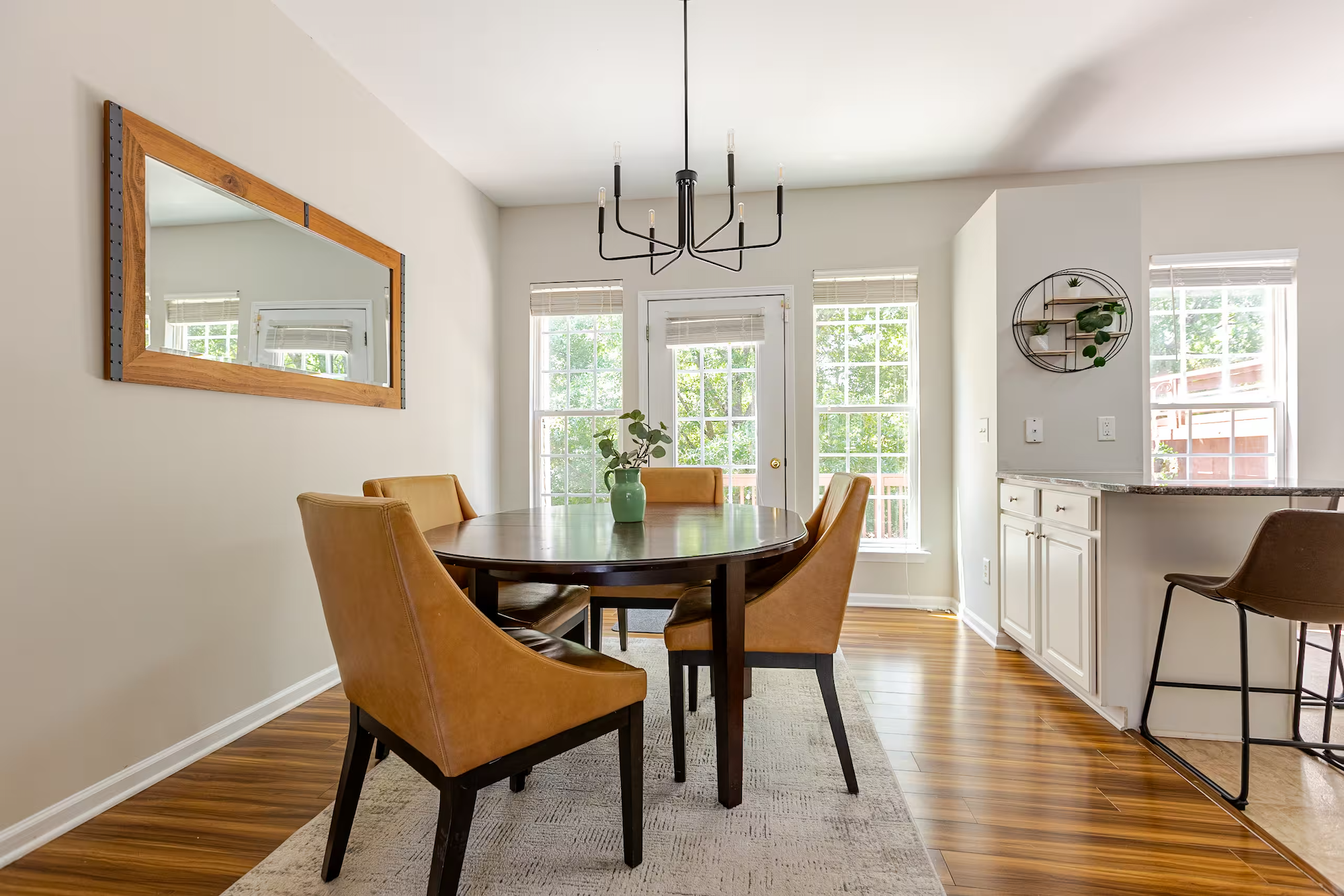 An inviting dining area of a Fort Collins STR with a wooden table and tan upholstered chairs, a black chandelier, large windows with blinds, a wall mirror, and a wall shelf with plants.