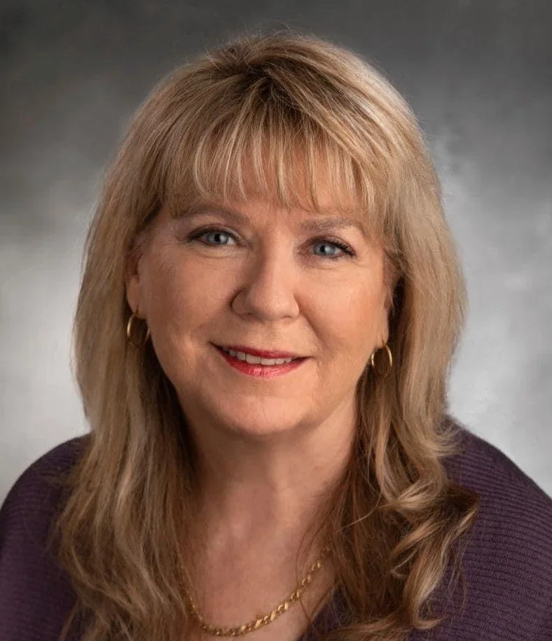 A portrait of a middle-aged woman with long, wavy blonde hair, blue eyes, and fair skin, smiling at the camera, wearing a dark purple top, gold hoop earrings, and a gold necklace.