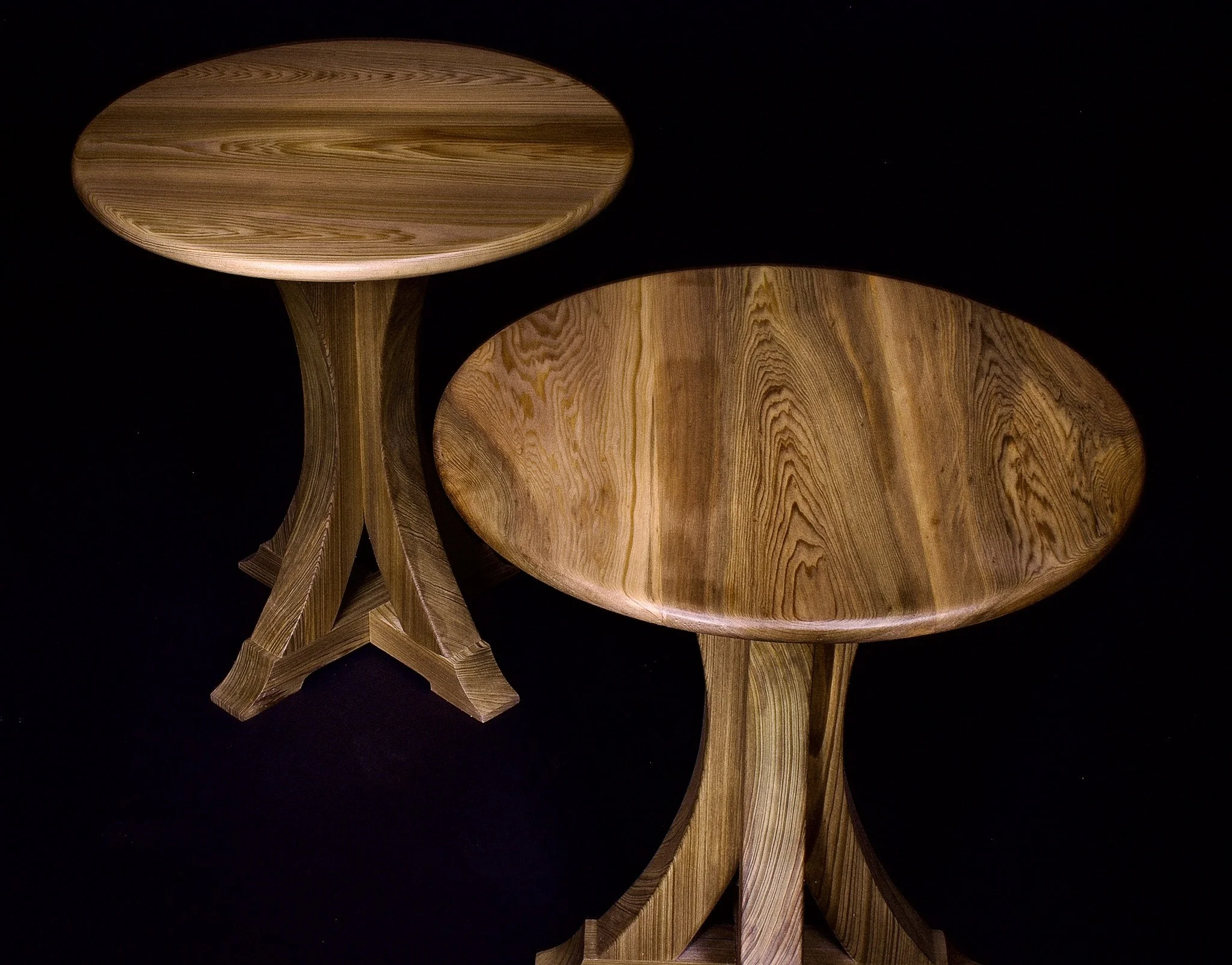 Two wooden end tables with oval tabletops and curved, supporting legs, set against a black background.