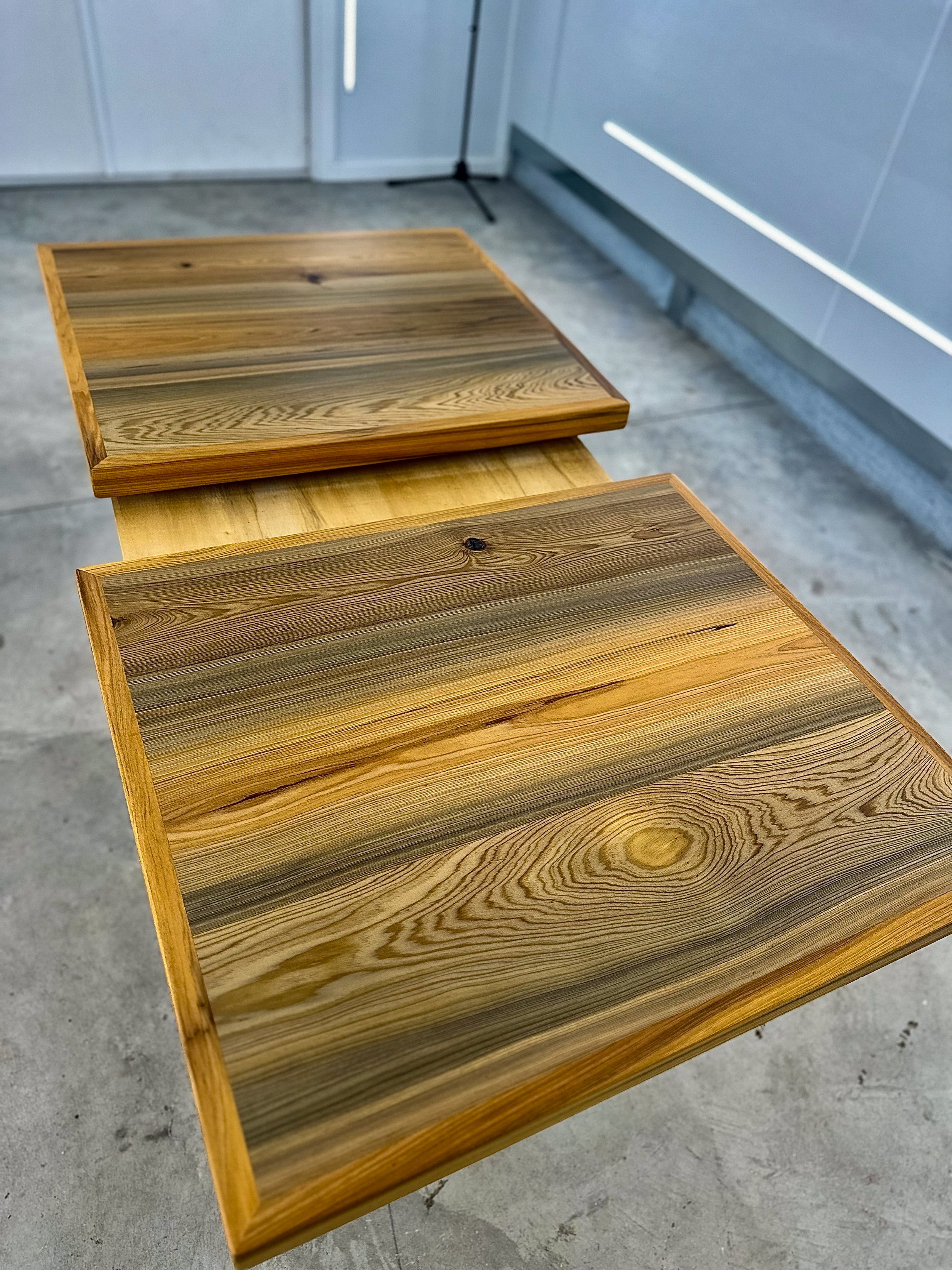 Two wooden tables with natural wood grain placed on a concrete floor near a door and wall.