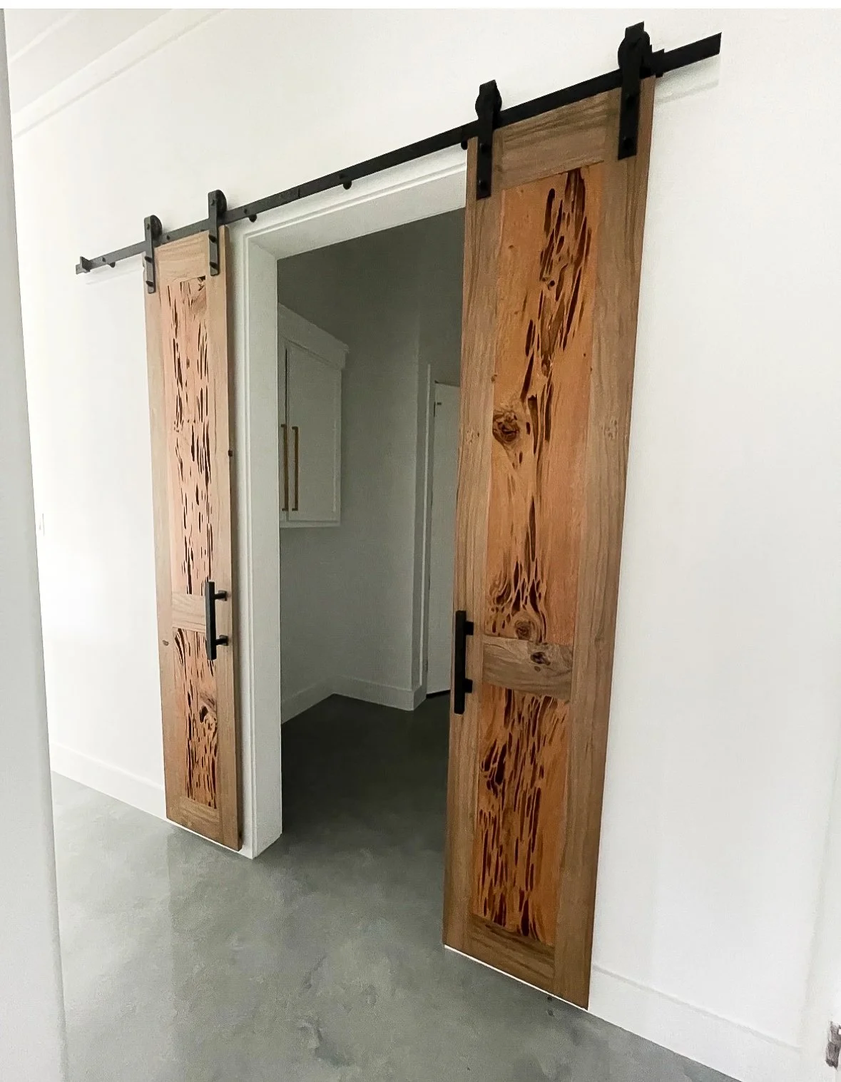Wooden sliding barn doors with black hardware, open to reveal a white room with white cabinets and gray flooring.