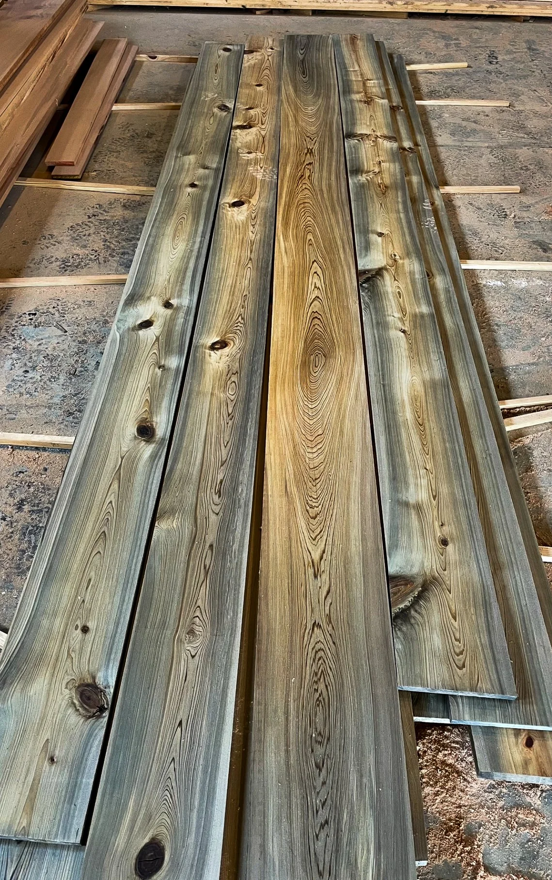 Several wooden planks with visible grain and knots laid out on a workshop floor, some supported by small wooden sticks.