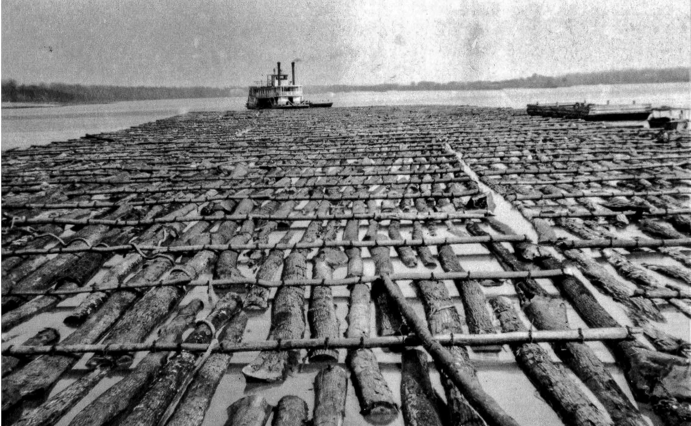 A black and white photograph of a makeshift raft made of logs and sticks tied together floating on a river. A small boat is visible in the background on the river.