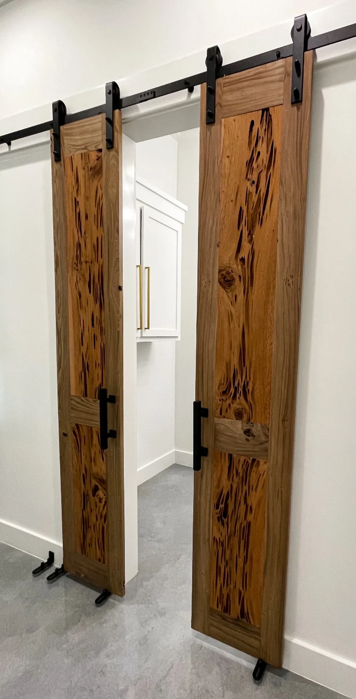 Wooden barn-style sliding doors with black hardware, partially open, revealing a white cabinet inside a room with white walls and gray flooring.