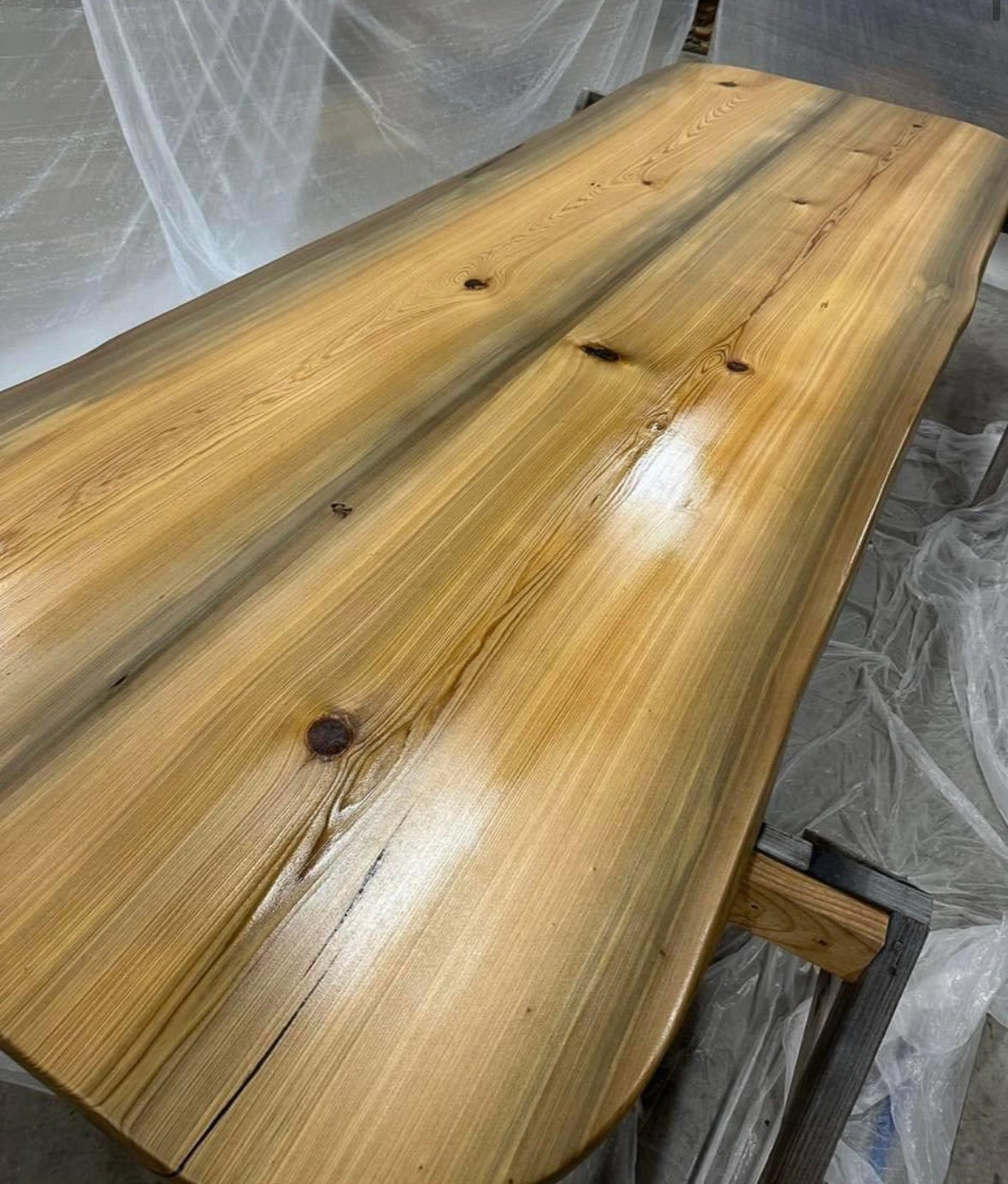 A polished wooden conference table with visible natural grain and knots and live edge, placed on a stand in a workshop setting with plastic sheeting in the background.