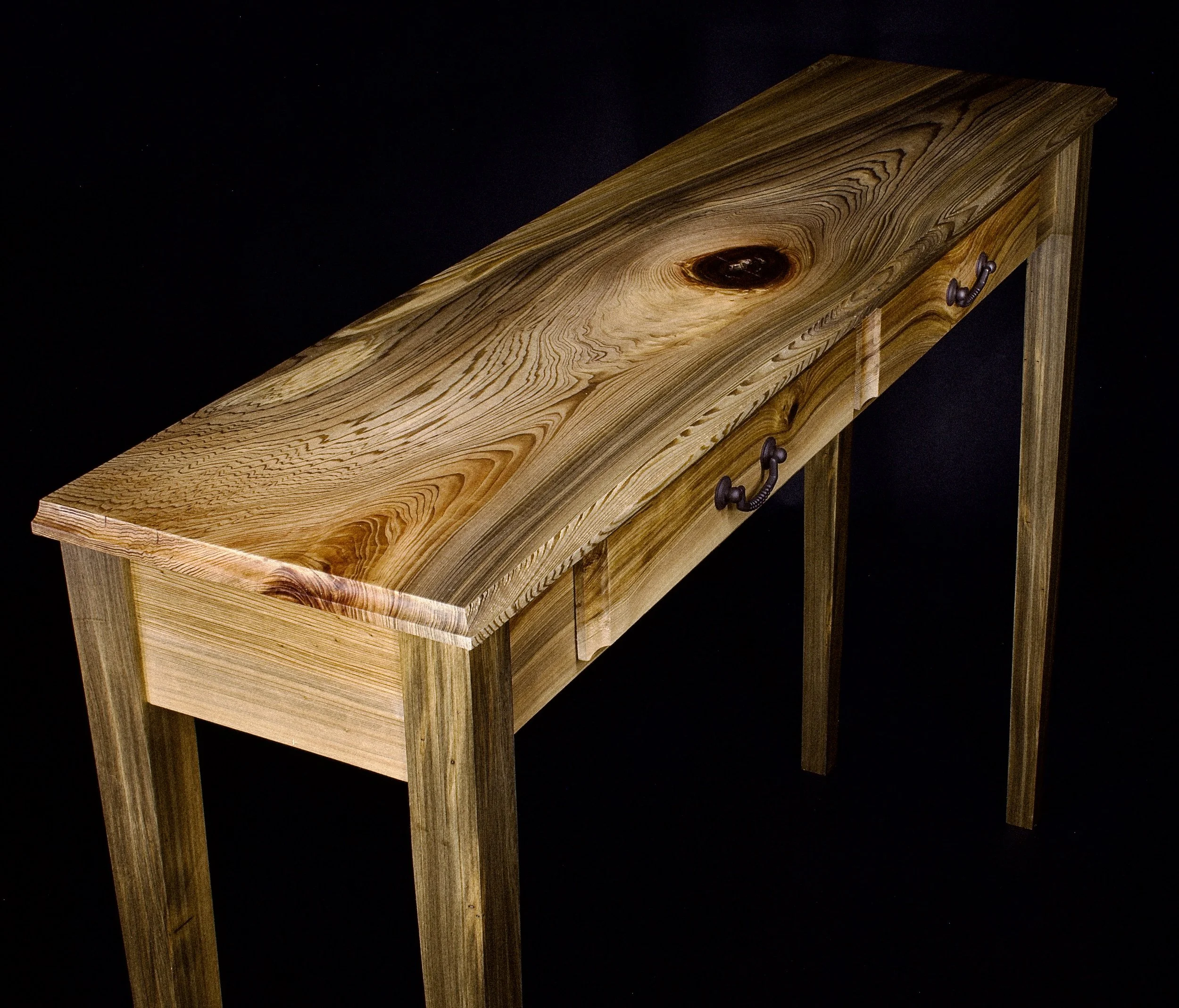 A wooden desk with a natural finish, featuring a wood grain top and metal handles on the drawers, set against a black background.